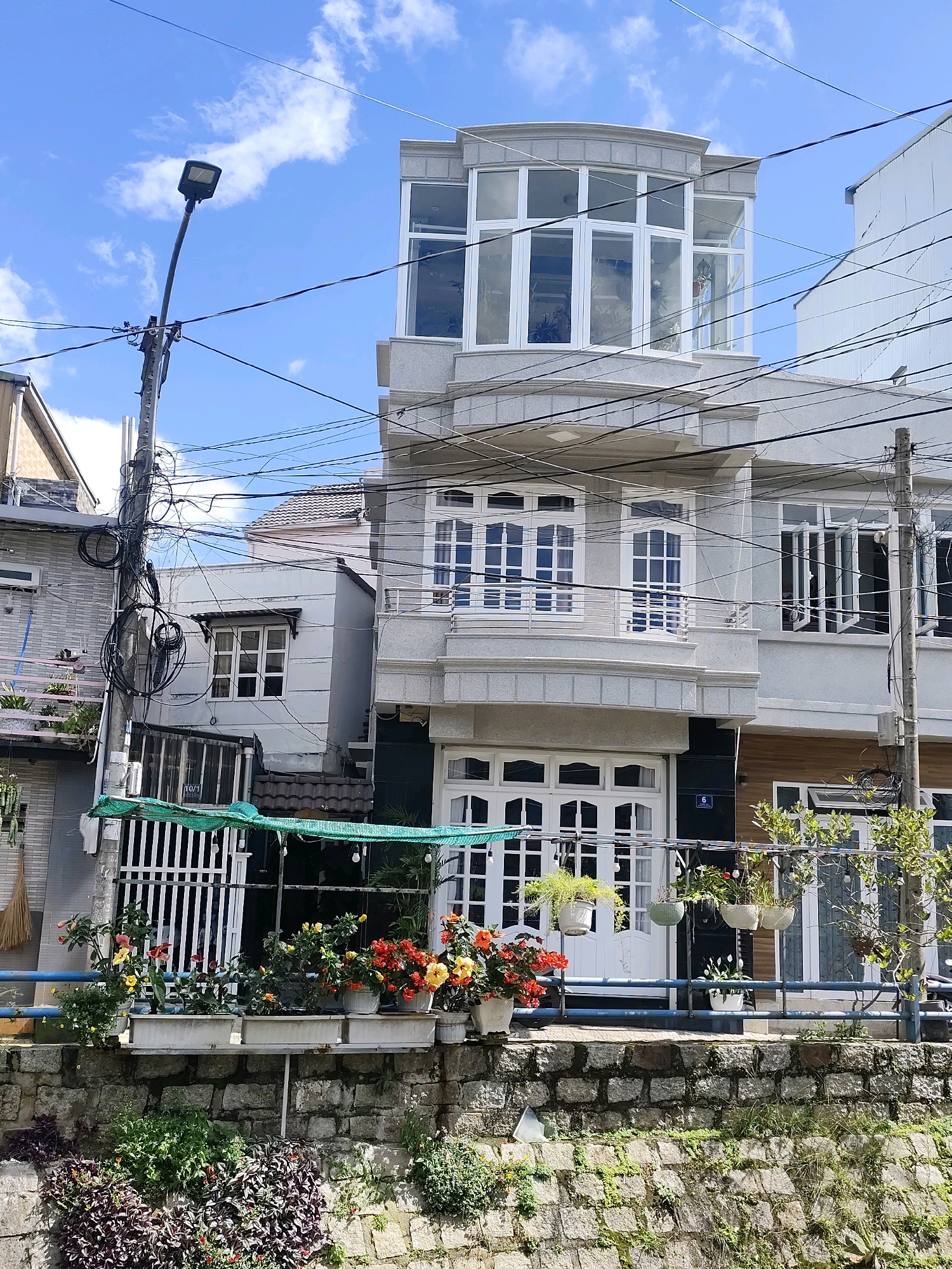 This image captures a multi-story building with a modern facade, likely in Da Lat, Vietnam, given the architectural style and lush greenery. The building features a prominent upper-level conservatory with large windows, characteristic of homes in the region that seek to maximize natural light. A street lamp stands to the left, its pole laden with a complex network of electrical wires, a common sight in Vietnamese urban landscapes. The building's exterior is predominantly gray, with white-framed windows and balconies.
In the foreground, a stone wall forms a retaining barrier, supporting a planter box filled with an array of colorful flowers, including red, yellow, and orange blooms. Below this, a dense collection of green and purple foliage covers the lower part of the wall. Hanging plants and more flowers in pots are visible on the building's balconies and near the entrance, adding a touch of vibrancy to the scene. The sky above is blue with scattered white clouds. The overall impression is of a residential building in a well-established urban area, characterized by both functional architecture and a love for decorative plant life.