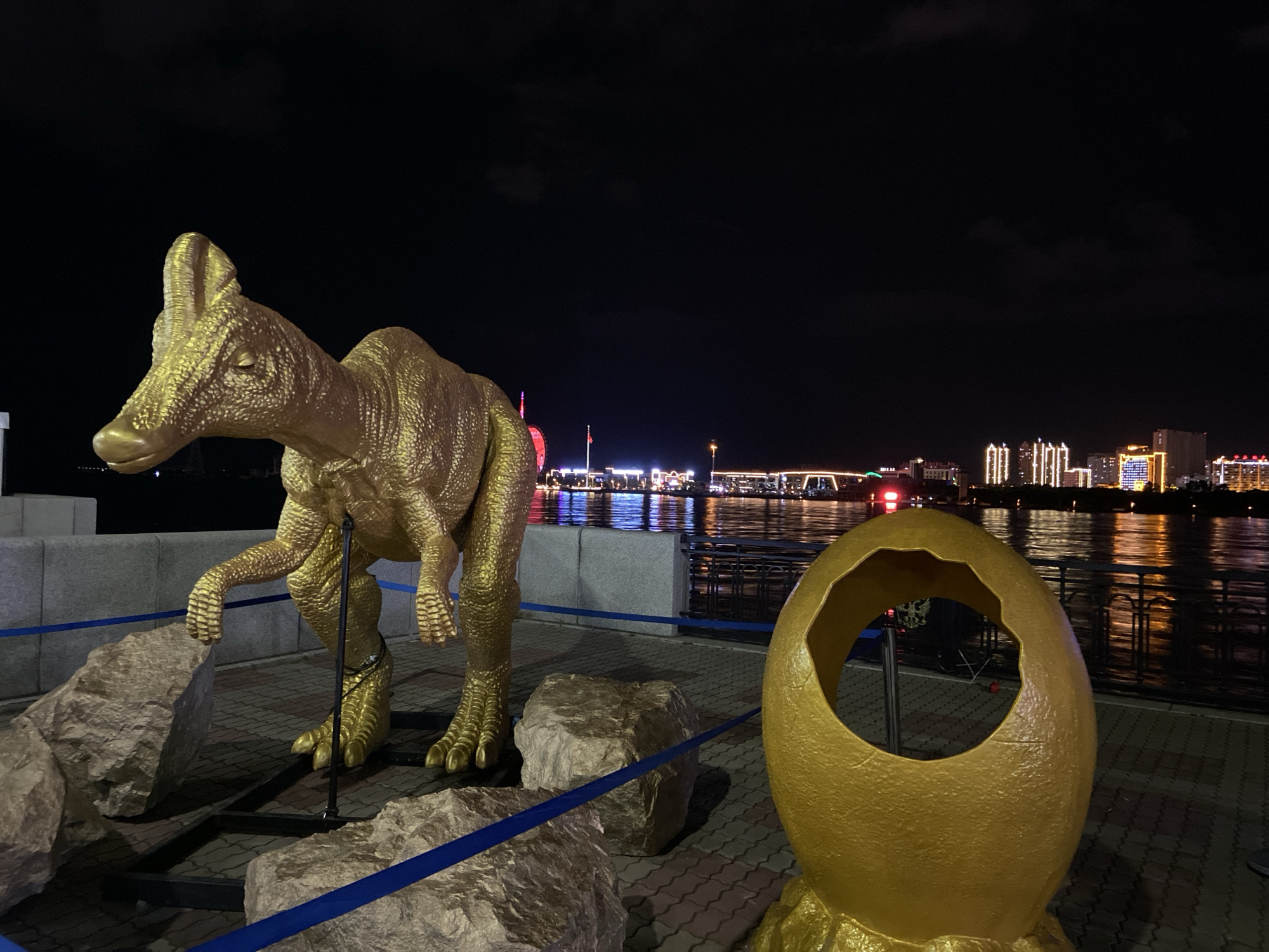 At night, a golden dinosaur statue stands on a paved waterfront promenade in Blagoveshchensk, Russia, adjacent to a large, golden, broken egg-shaped sculpture. The dinosaur, which appears to be a Parasaurolophus, is positioned to the left of the frame, with its head turned slightly to the left, and its front right leg extended forward. Several large, grey rocks are placed around the base of the dinosaur. Blue velvet ropes cordon off the display. In the background, the Amur River flows, and across the river, the city lights of China's Heihe are visible, reflecting on the water. The sky is dark and clear, with a few faint clouds. There are no people visible in the image.