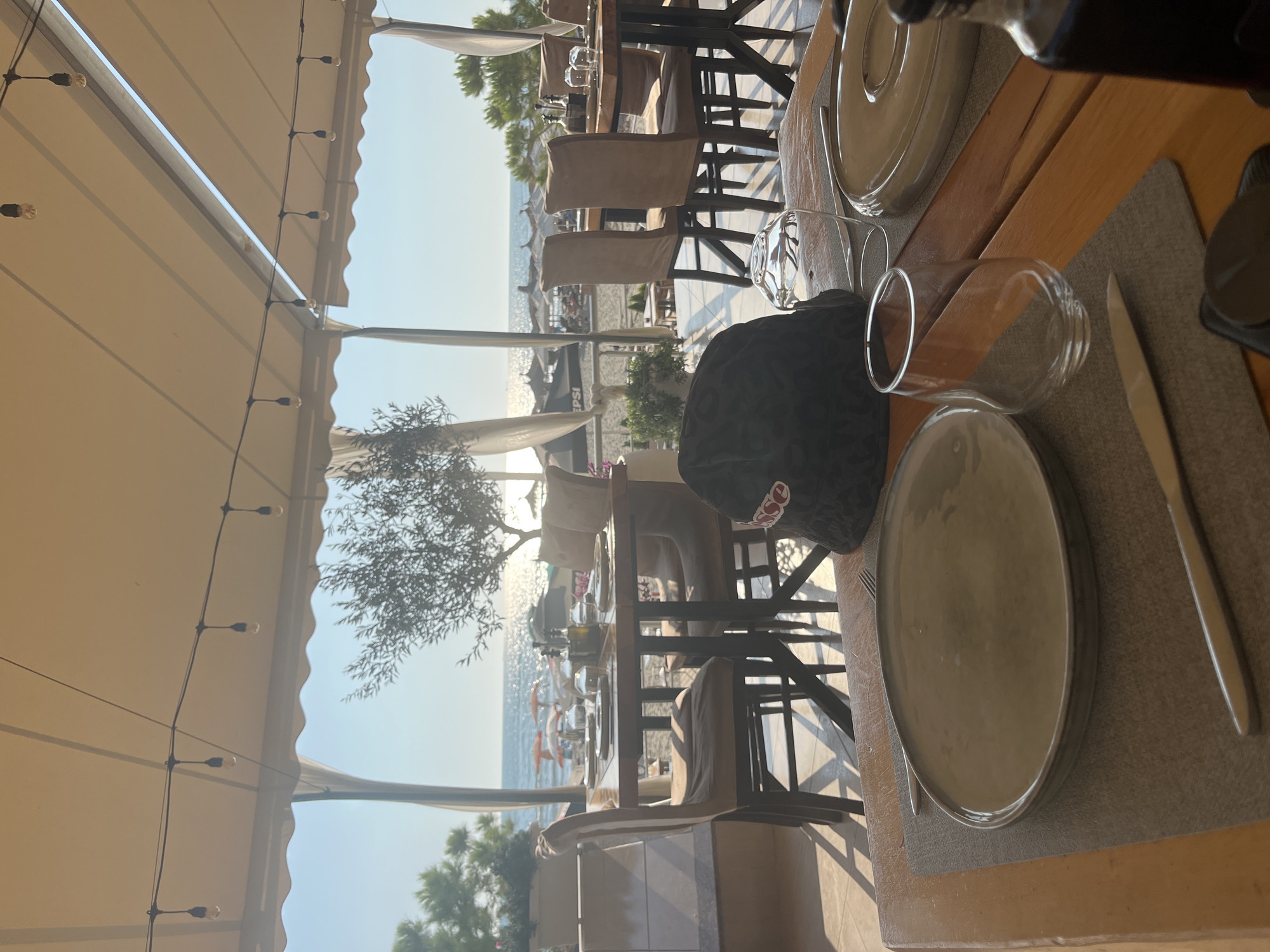 The image depicts an outdoor dining area of a restaurant located in Bashkia Durrës, Albania, with a view of the beach and the Adriatic Sea. The scene is set during daylight, with sunlight filtering through an awning. In the foreground, a wooden table is set with place settings including plates, cutlery, and glasses. A dark-colored baseball cap with the word "EPSO" is placed on the table. Beyond the table, several chairs are arranged around other dining tables. In the background, the beach is visible with numerous umbrellas and people. The sea stretches to the horizon under a clear sky. The overall atmosphere suggests a relaxed seaside dining experience.