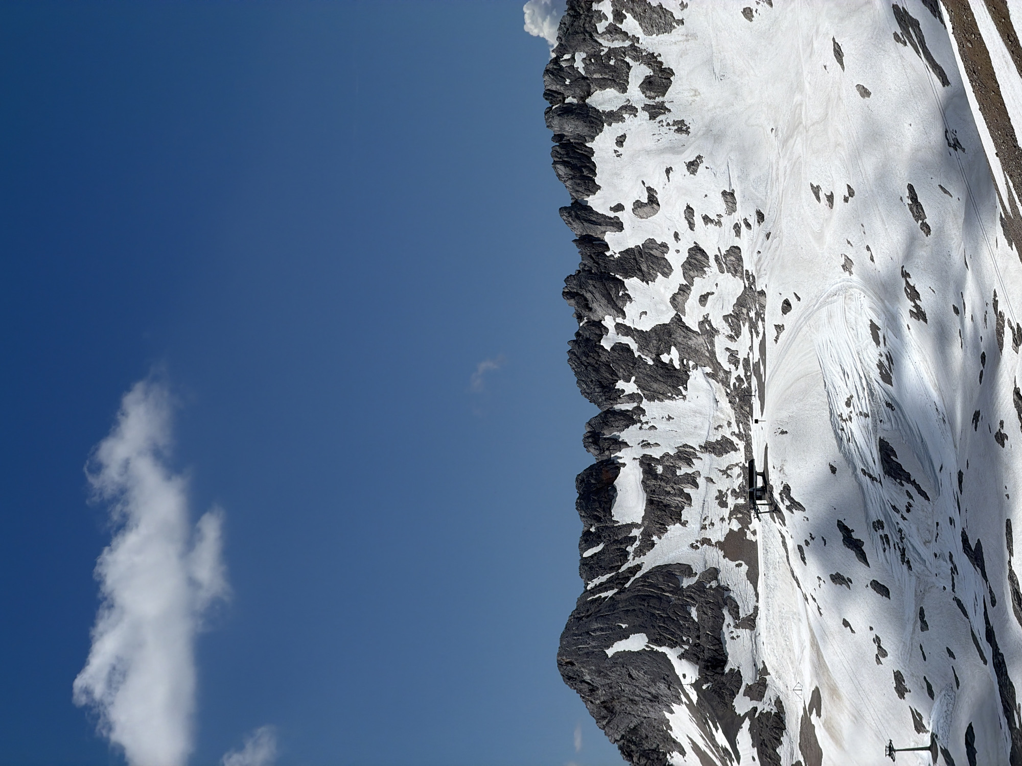 The image shows a panoramic view of the Zugspitze mountain range near Garmisch-Partenkirchen, Germany. The upper portion of the image is dominated by a clear blue sky with a single white cloud. The lower portion features jagged, dark grey mountain peaks partially covered in snow. A vast, snow-covered slope extends from the base of the peaks, with patches of dark rock and soil showing through the snow. There are ski tracks visible on the slope, indicating it's a ski area. A ski lift station with a dark roof and supporting structures is situated on the slope, along with a ski lift tower to the left. The overall environment is alpine, characterized by mountains, snow, and a clear sky, suggesting a high altitude location during a fair-weather day.
