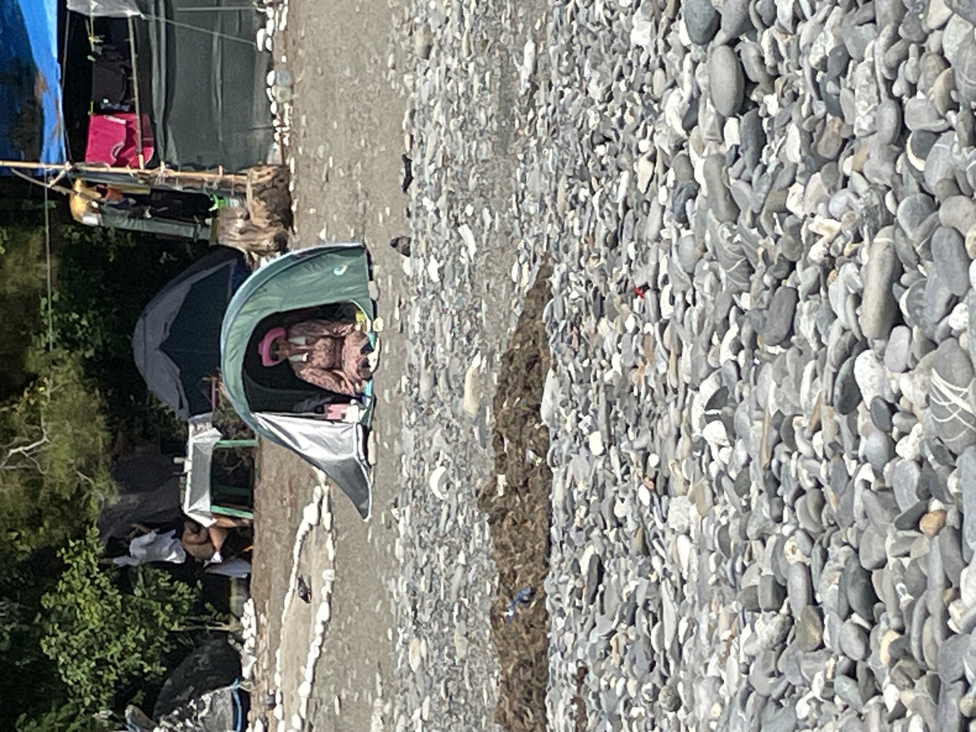 The image depicts a scene on a pebble beach, possibly in Sochi, Russia, given the context. In the foreground, a person wearing a pink hat and a patterned dress is seated inside a green tent. The tent is open, allowing the person to be partially visible. The beach is covered in smooth, grey pebbles of various sizes.
In the mid-ground, there are more tents and a table set up, with some figures in the background. One person in a white shirt appears to be interacting with someone seated near the table. The background shows trees and vegetation, suggesting a natural setting adjacent to the beach.
Further back, a larger tent structure is visible, with clothing hung on a pole. The overall impression is of a temporary encampment or a group of people spending time outdoors on the coast. The lighting suggests it is daytime, with the sun casting shadows.