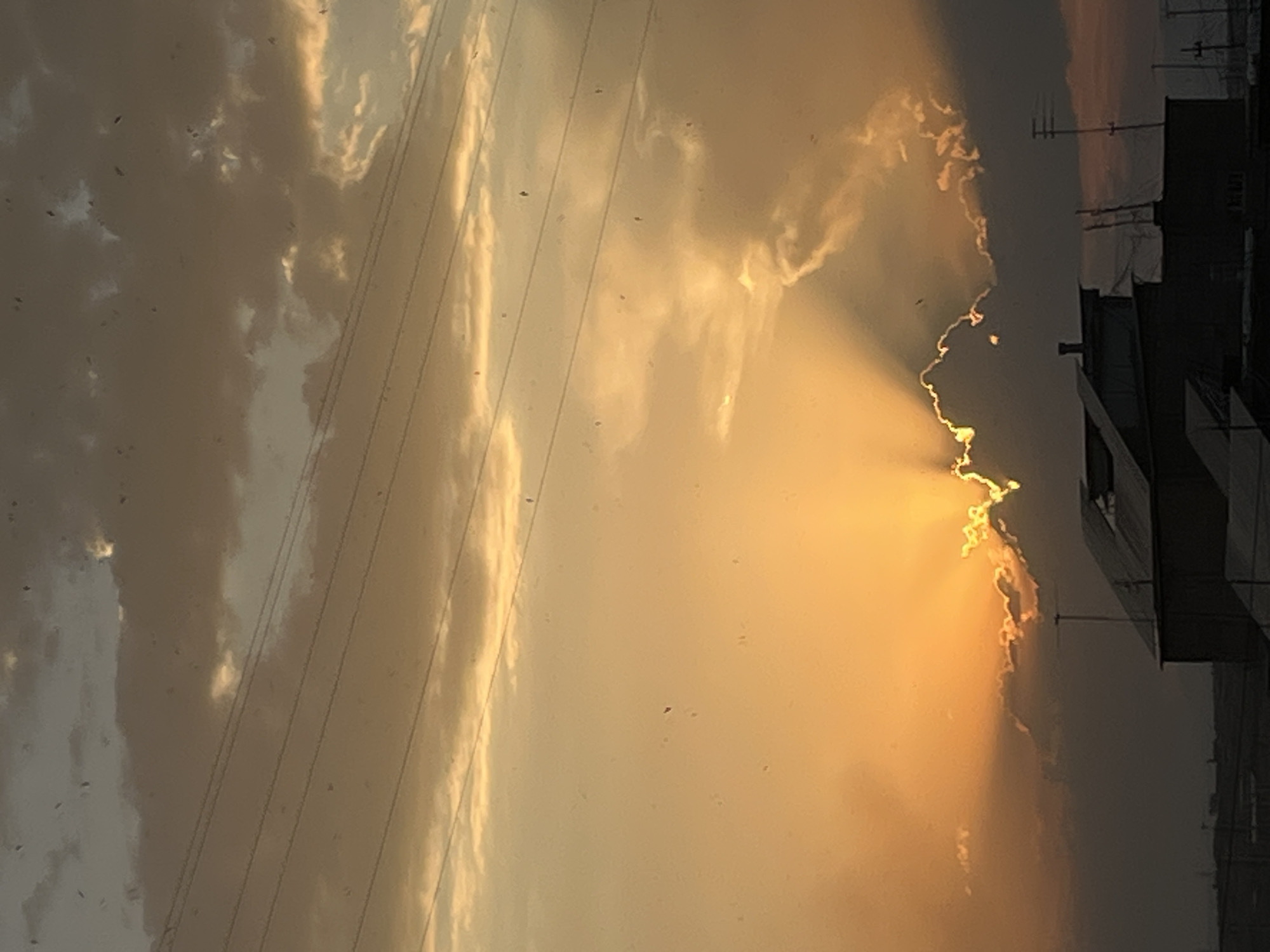 This image captures a dramatic sunset over Yerevan, Armenia. The sun, partially obscured by dark clouds, casts intense golden rays that fan out across the sky. These beams of light highlight the edges of the clouds, creating a vibrant contrast between light and shadow. In the foreground, the silhouettes of buildings and rooftops are visible against the illuminated sky, with a few antennas and utility poles also dotting the landscape. The overall impression is one of a striking natural phenomenon occurring within an urban setting.