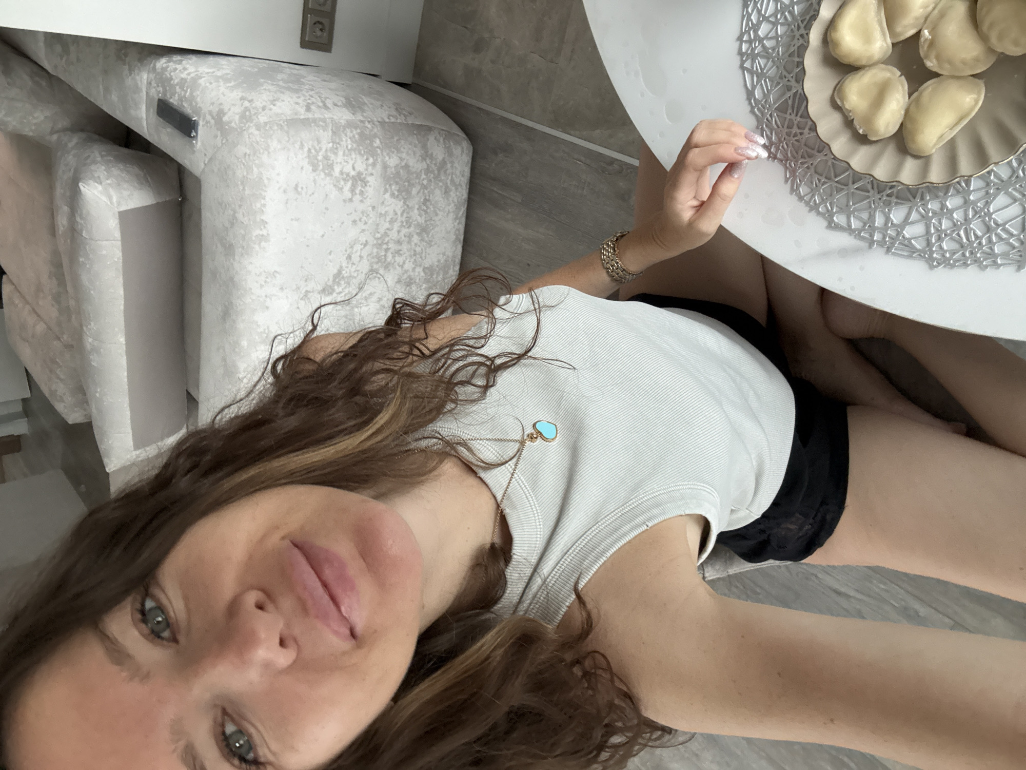 A woman is sitting at a white round table in a well-lit room that appears to be a living room in Khimki, Russia. The woman is taking a selfie with her right hand, and her face is close to the camera. She has long, wavy brown hair and green eyes. She is wearing a white tank top and black shorts. Around her neck, she has a gold necklace with a turquoise heart-shaped pendant. On her left wrist, she is wearing a gold bracelet. To her right, on the table, is a plate with several dumplings on it. In the background, a large, modern sectional sofa with a textured white fabric is visible. The flooring appears to be laminate or tile. Electrical outlets are visible on the wall in the background.