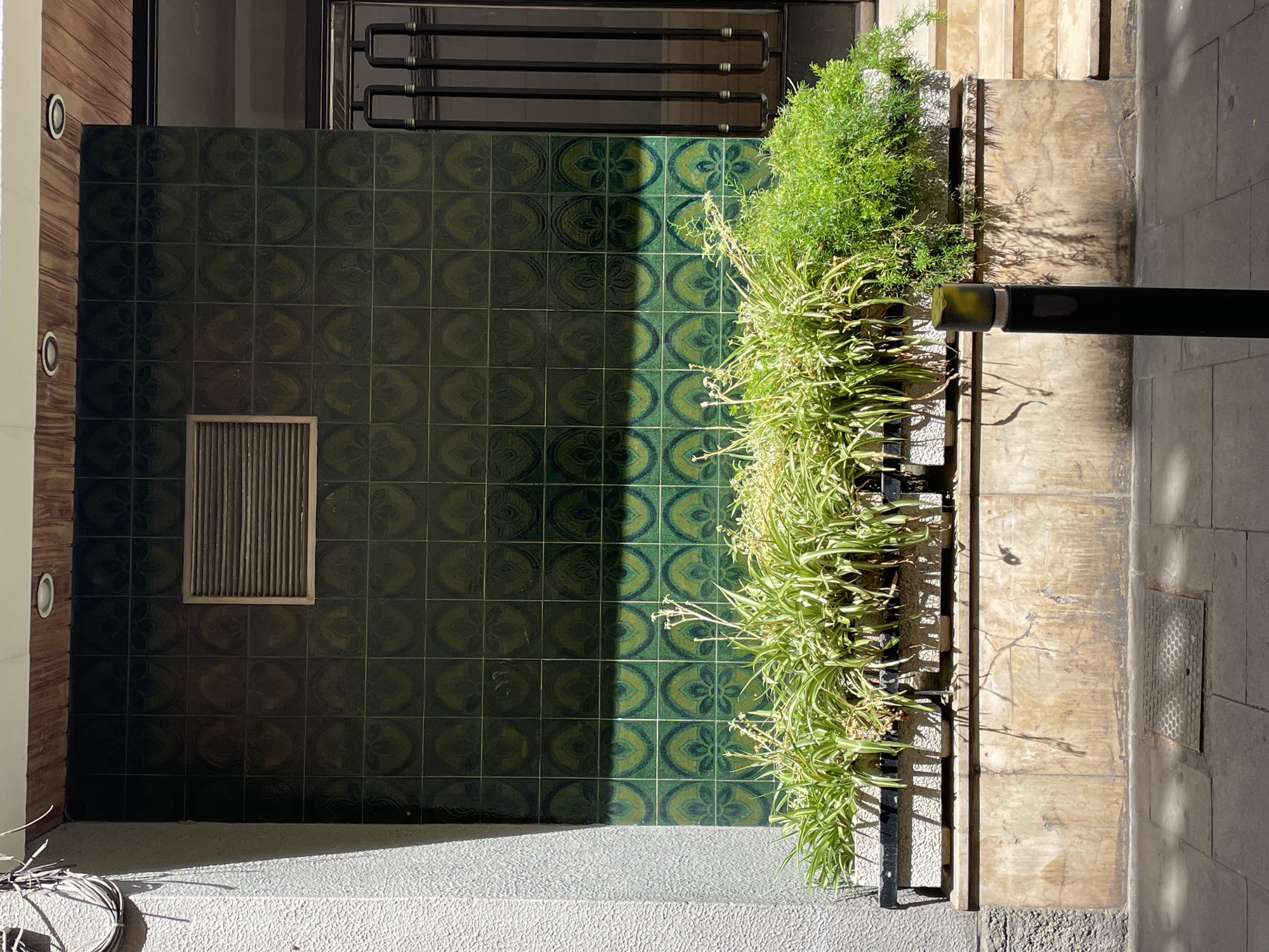 This is a frontal, eye-level shot of a building facade in Barcelona, Spain, featuring green tiles with a circular pattern, a ventilation grate, planters with greenery, and a sidewalk. The building has a textured gray wall on the left and a dark door on the right. The ground level is composed of beige stone panels. Sunlight casts shadows on the facade and the sidewalk. A black bollard with a yellow top stands in front of the planters. The scene appears to be a street-level view, possibly in a residential or commercial area. There are no people present.
