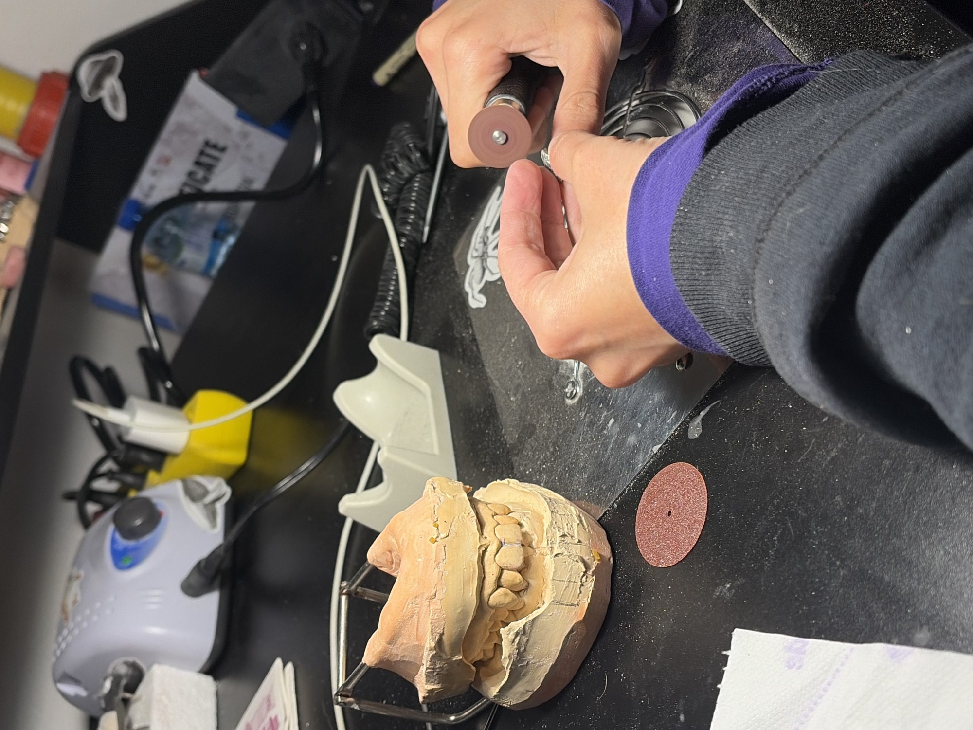 The image depicts a dental technician at work, potentially in a dental lab, but location specifics for Sofia, Bulgaria, are unavailable. A person is shown holding a small rotary tool with a grinding disc, appearing to be working on a small object. To the lower left, there is a dental model attached to a metal articulator, along with a small machine with dials and buttons, potentially a dental drill or polisher. A second grinding disc is on the table next to the model. The work surface is black and has several cables and tools nearby.