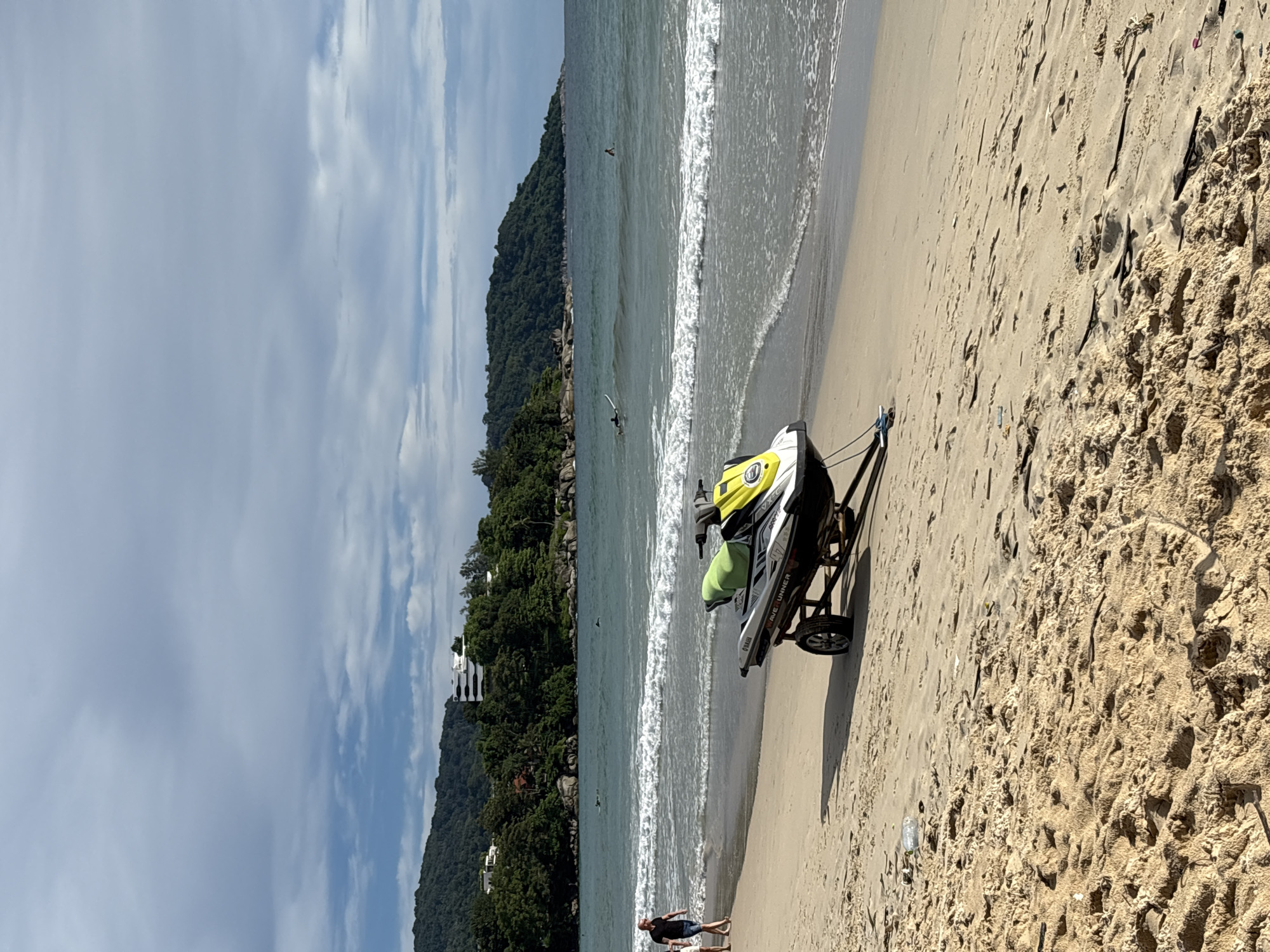A jet ski is parked on a sandy beach in Karon, Thailand. A person is walking on the beach in the background, and there are waves in the ocean. The sky is partly cloudy. The jet ski is yellow and white and is on a trailer. There are trees and hills in the background.