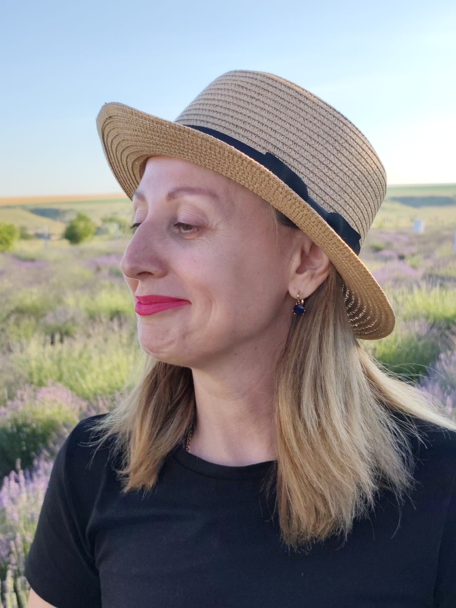 This is a medium shot portrait of a woman wearing a straw hat and a black t-shirt, taken outdoors. She is looking to her left, with her head slightly tilted. Her eyes are closed and she has bright pink lipstick on. She has blonde hair that is visible from under her hat. She is wearing a small dark earring in her visible ear.
The background appears to be a field of lavender under a clear blue sky, suggesting it is daytime and likely warm weather. The lighting indicates it might be late afternoon or early morning due to the soft light. The overall mood seems peaceful and serene.
The woman's expression, with her eyes closed and a slight smile, conveys a sense of contentment or enjoyment of the moment. The setting of a lavender field in bloom adds to this peaceful and idyllic atmosphere.