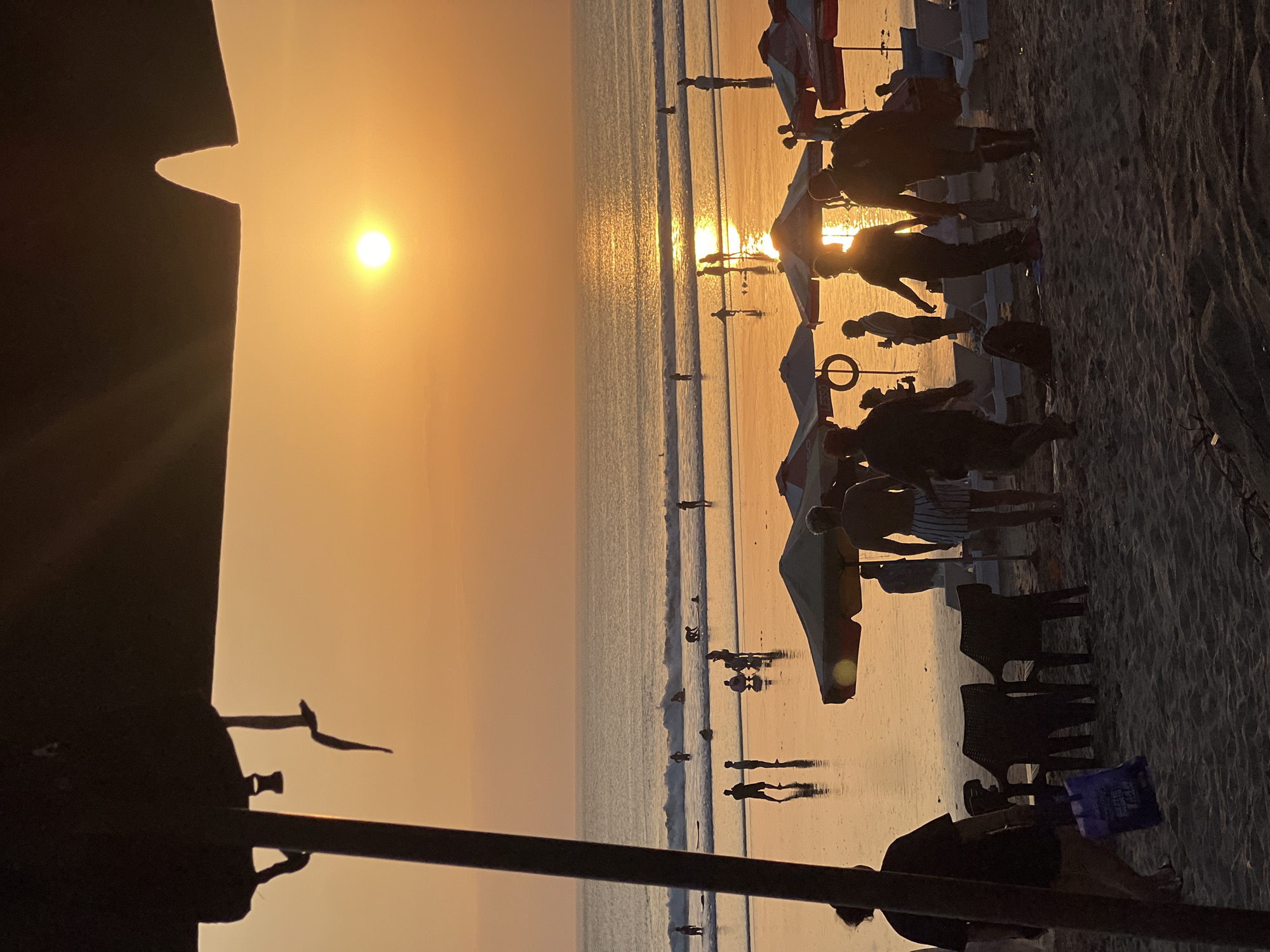 This image depicts a sunset scene on Kuta Beach, Bali, Indonesia. The sun is low in the sky, casting a warm, orange glow across the water and sand. The ocean is calm with small waves, and several people are visible in the water, some appearing to be swimming or wading. On the wet sand near the water's edge, more figures are scattered, some interacting with each other. Beach umbrellas and lounge chairs are set up on the drier sand further back, with several silhouetted individuals standing or walking among them. A dark, silhouetted structure, likely a part of a beach umbrella or awning, is prominent in the foreground on the left, partially obscuring the view of the beach and sky. The overall atmosphere is tranquil and evokes a typical beach evening.