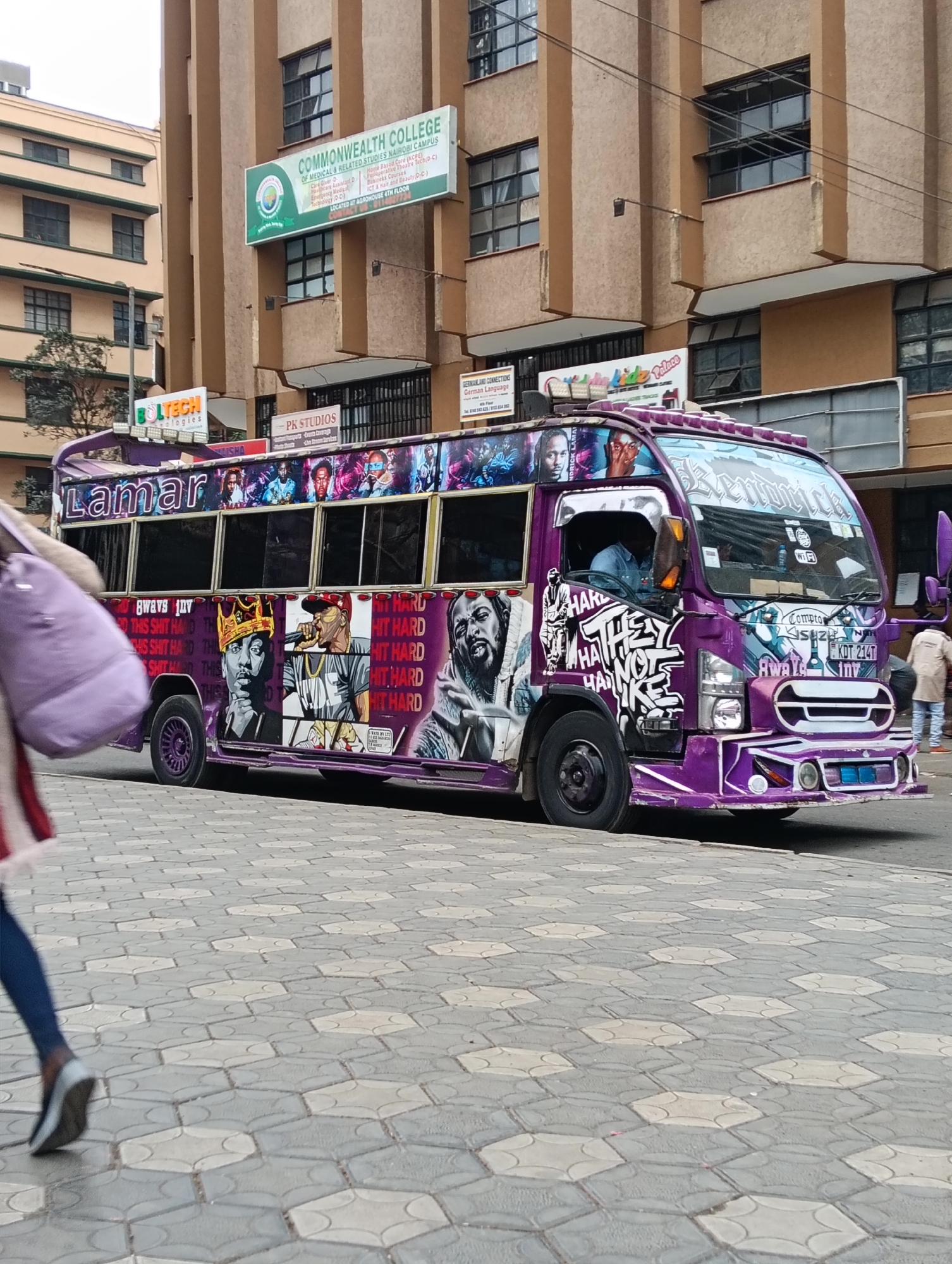 The image captures a vibrant purple matatu, a form of public transportation common in Nairobi, Kenya, parked on a street. The matatu is heavily decorated with graffiti and portraits of various figures, including what appear to be rappers and musicians. Text on the side of the vehicle reads "Lamar" and other slogans. The matatu has tinted windows and is adorned with numerous lights and artistic details.
In the background, a multi-story building with a beige facade and dark-rimmed windows stands prominently. A sign on the building reads "COMMONWEALTH COLLEGE OF MEDICAL & RELATED STUDIES NAIROBI CAMPUS". Another sign with the text "PK STUDIOS" is also visible. The street is paved with hexagonal interlocking concrete blocks, typical of Nairobi's urban landscape.
In the foreground, a person is walking past the matatu, their legs and lower body visible as they move from left to right. The person is wearing blue leggings and grey shoes, and a purple bag is slung over their shoulder. The scene suggests a bustling urban environment in Nairobi, Kenya, characterized by the distinctive local matatu culture and the presence of educational institutions. The overall impression is one of a lively street scene with a focus on the artistically decorated public transport.