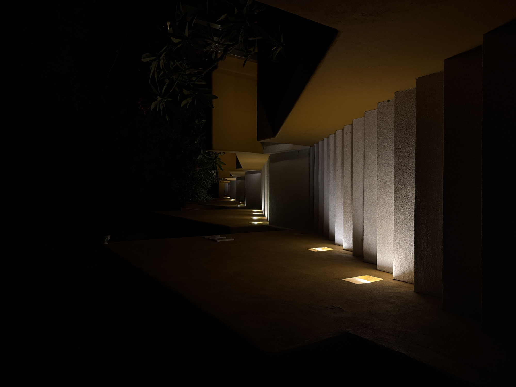 This is a nighttime scene in Puerto Marqués, Mexico, featuring a partially illuminated walkway. The walkway is flanked by a series of vertical concrete structures or pillars that are lit from below by embedded, square-shaped lights. The pillars extend towards an overhanging structure, possibly a building or covered passageway, which casts a large shadow. Further in the distance, the walkway continues with additional in-ground lights illuminating the path. To the left, there's a dark area with vegetation or foliage, blending into the darkness. The foreground of the image is mostly dark. No people are visible. The overall impression is that of an outdoor architectural space with intentional lighting for ambiance and guidance.