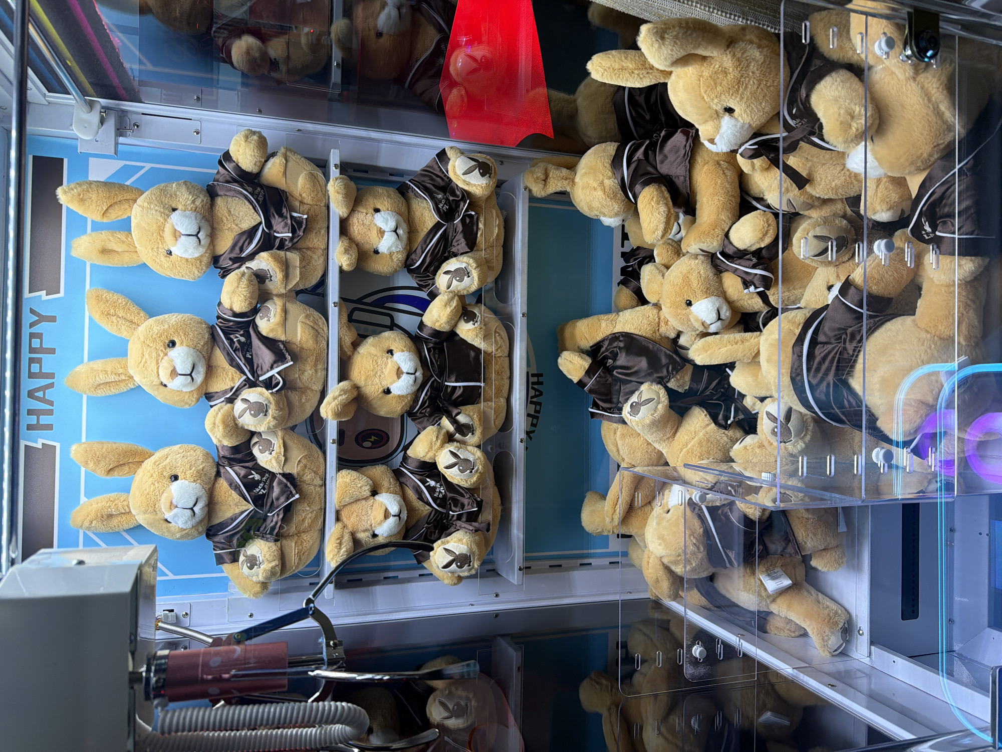 The image shows an inside view of a claw machine filled with plush bunny toys. The bunnies are light brown with white faces and wear brown jackets with a white logo and trim. The claw machine has a light blue interior wall with the word "HAPPY" printed on it. The metal claw arm and the inner workings of the machine are visible in the foreground. The reflection in the glass of the machine suggests an indoor environment. Based on the logos and branding on the plush toys, it is likely located in West Hollywood, United States of America.