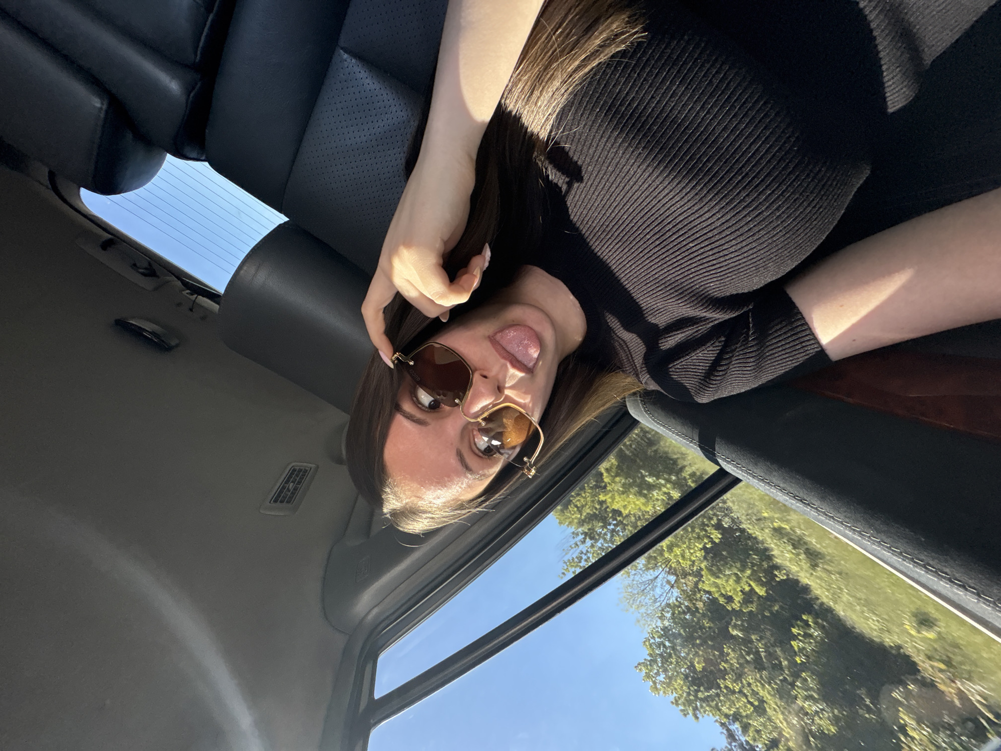 A person with long brown hair, wearing sunglasses and a black ribbed shirt, is sticking their tongue out. The person is inside a car with a sunroof, with trees and a blue sky visible through the windows. The car has black leather seats. There is no evidence of the location being Burgas, Bulgaria.