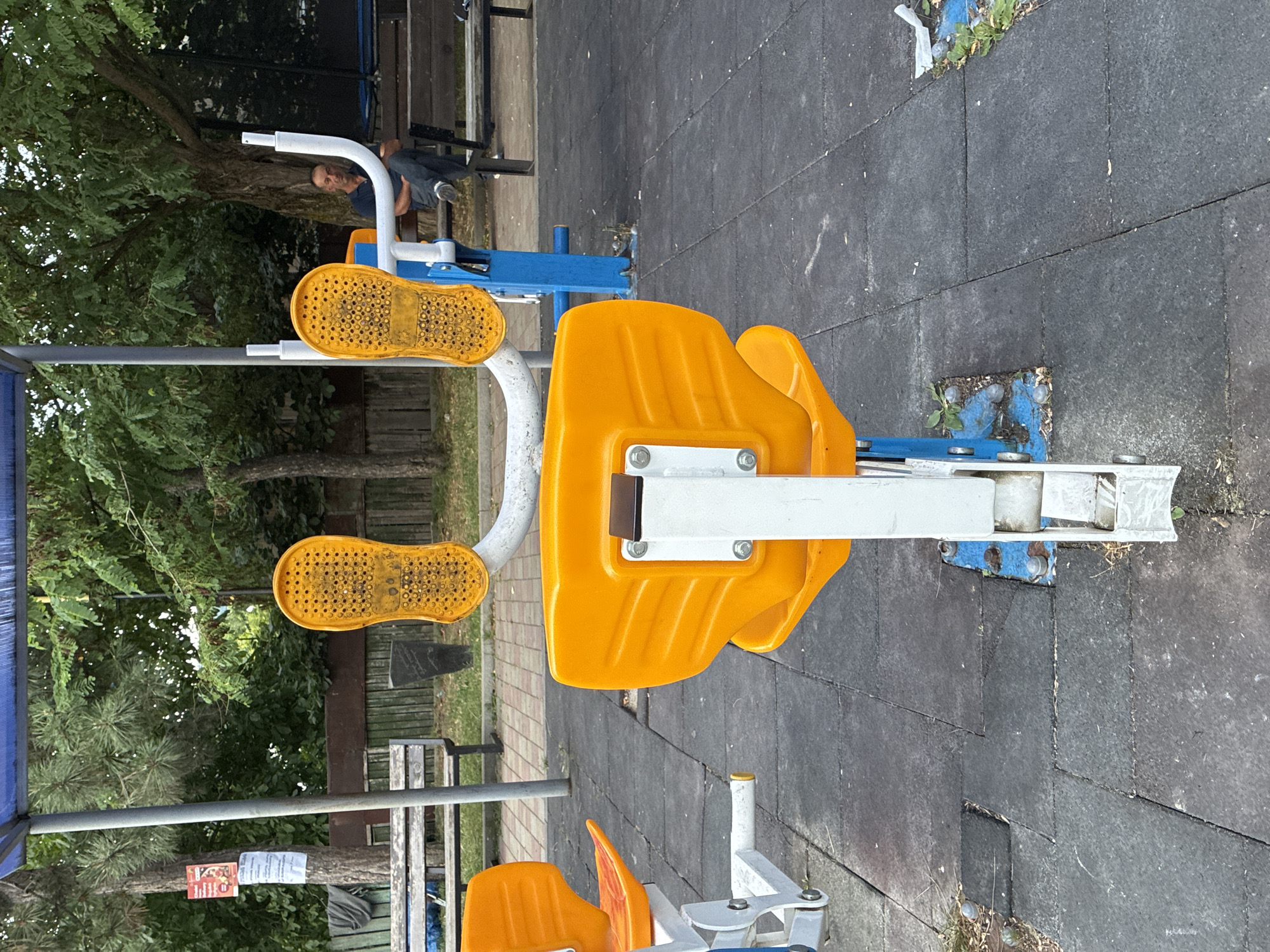 In Surami, Georgia, an outdoor exercise machine with bright orange seats and yellow footplates stands on a rubberized playground surface. The machine is positioned under a blue canopy structure, with metal poles supporting it. Behind the machine, a man is seated on a wooden bench, looking to his left. Trees and foliage surround the area, and a fence can be seen in the background.