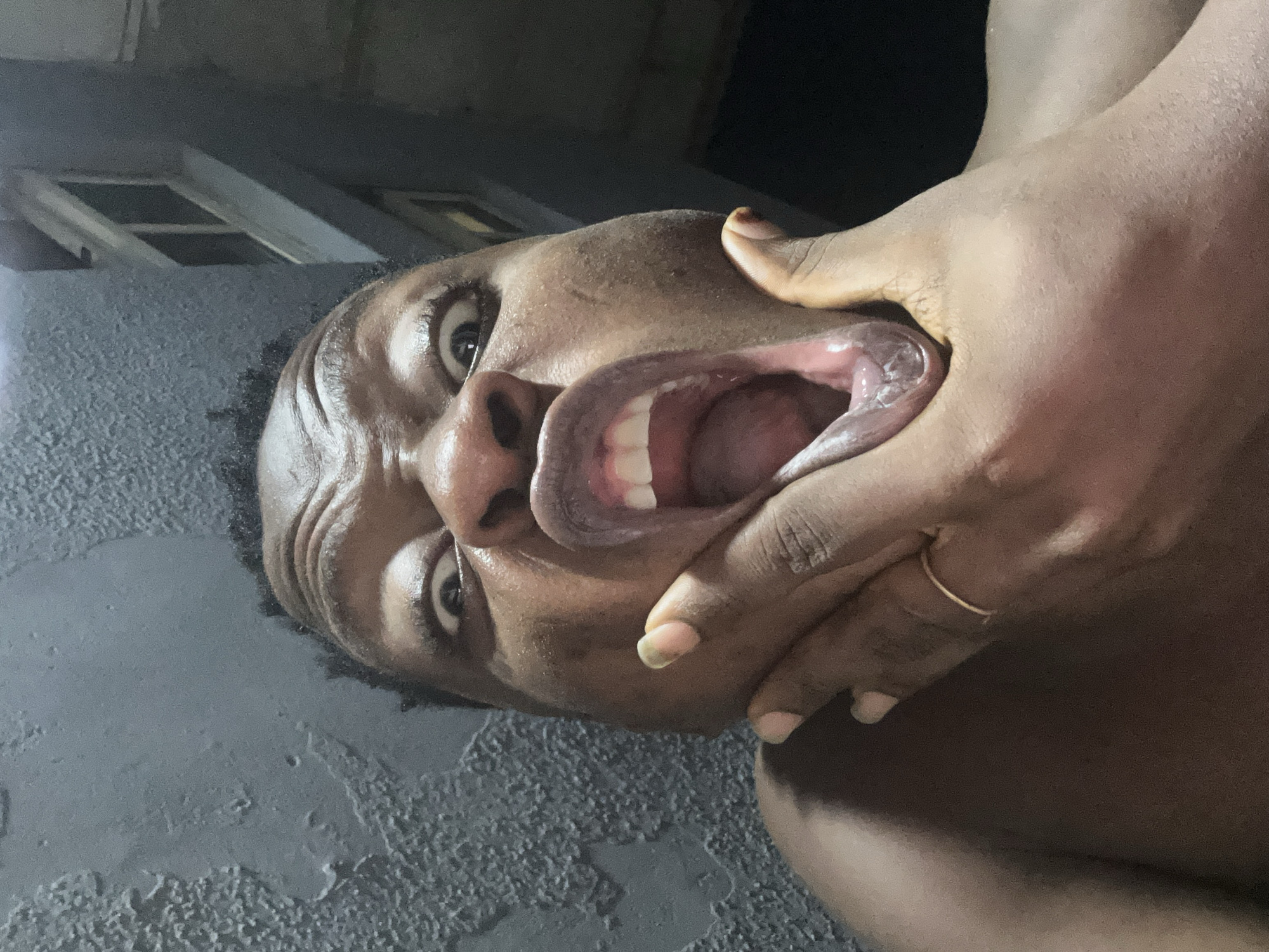The image presents a close-up portrait of a single individual, an adult with dark skin, captured in an exaggerated pose. The person's face is central, with eyes wide open and brows furrowed. The mouth is stretched wide, revealing the upper teeth and tongue. A hand, appearing to be the subject's left hand, is positioned to the lower left of the face, pulling the cheek and jaw downwards, thus distorting the mouth further. A thin, gold-colored ring is visible on a finger of this hand. The background on the left features a dark grey, unevenly textured surface, possibly a wall, with some lighter grey patches. The background on the right is darker and less distinct, showing rectilinear architectural features consistent with windows or structural elements of a building. The overall scene is dimly lit. The image contains no visual identifiers specific to the city of Ikorodu, Nigeria.