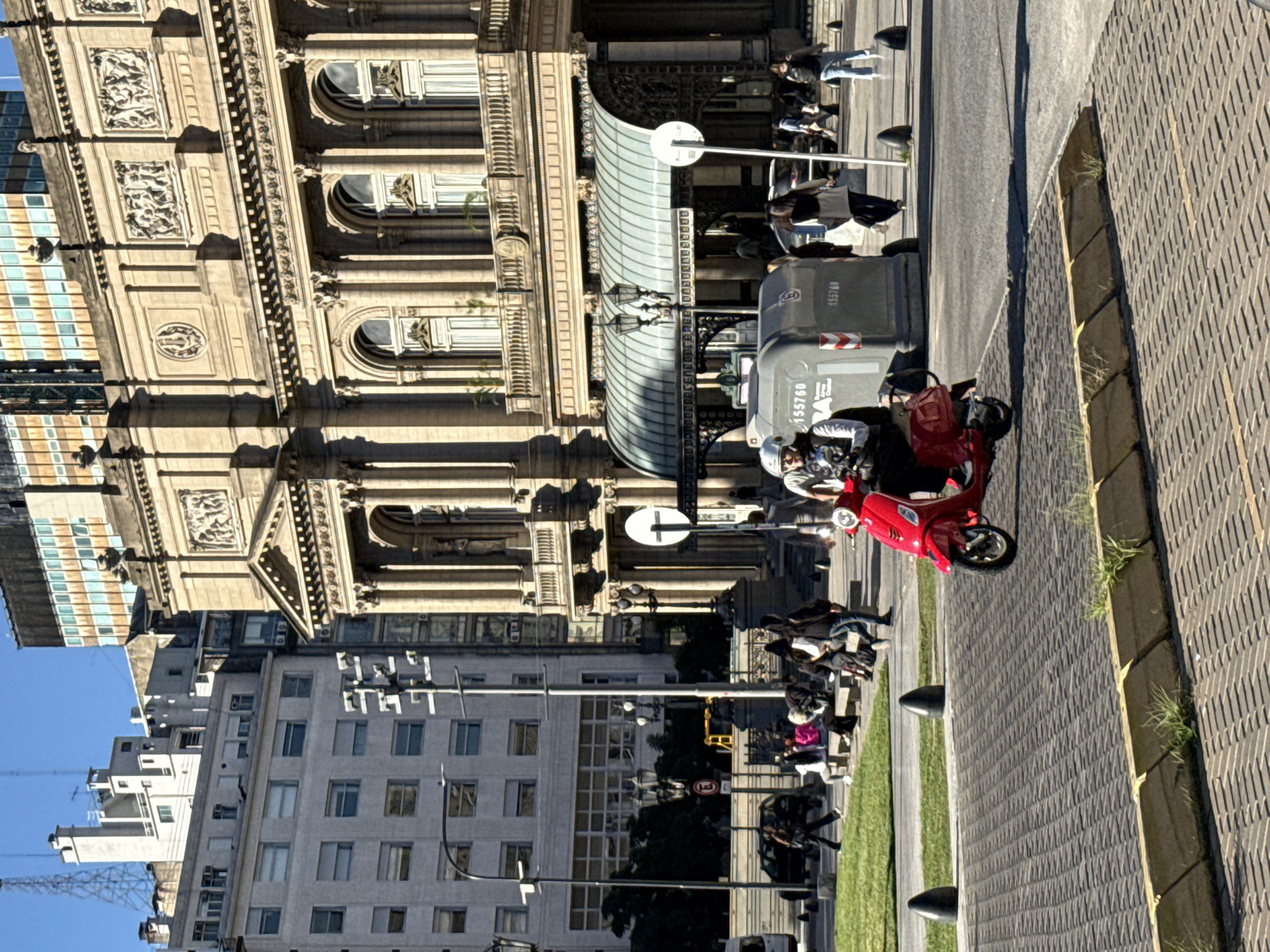 This image captures a vibrant street scene in Buenos Aires, Argentina, under a bright, clear blue sky. The photo is oriented ninety degrees clockwise.
In the foreground, a prominent red scooter is seen, with two individuals aboard, both wearing helmets and light-colored outerwear. The ground consists of a textured, patterned paving, possibly cobblestones or blocks, alongside a strip of manicured grass and black bollards.
The midground features a street with pedestrians and vehicles. A large grey municipal garbage bin is clearly visible, bearing the identification number "155760" twice, along with a red and white chevron sticker. A white circular sign on a pole stands nearby, typically indicating a traffic instruction. People are seen walking and congregating on sidewalks.
The background showcases a blend of architectural styles. A grand, ornate building with classical detailing, including intricate carvings, columns, and arched windows, dominates the left side. Beneath it, a curved glass and metal canopy extends over an entrance. Adjacent to and behind this historic structure, more modern multi-story buildings with numerous windows rise, reflecting the blue sky. A construction crane is discernible in the distant skyline. The overall scene depicts a sunny, busy daytime in an urban setting, likely a historical or commercial area of the city.