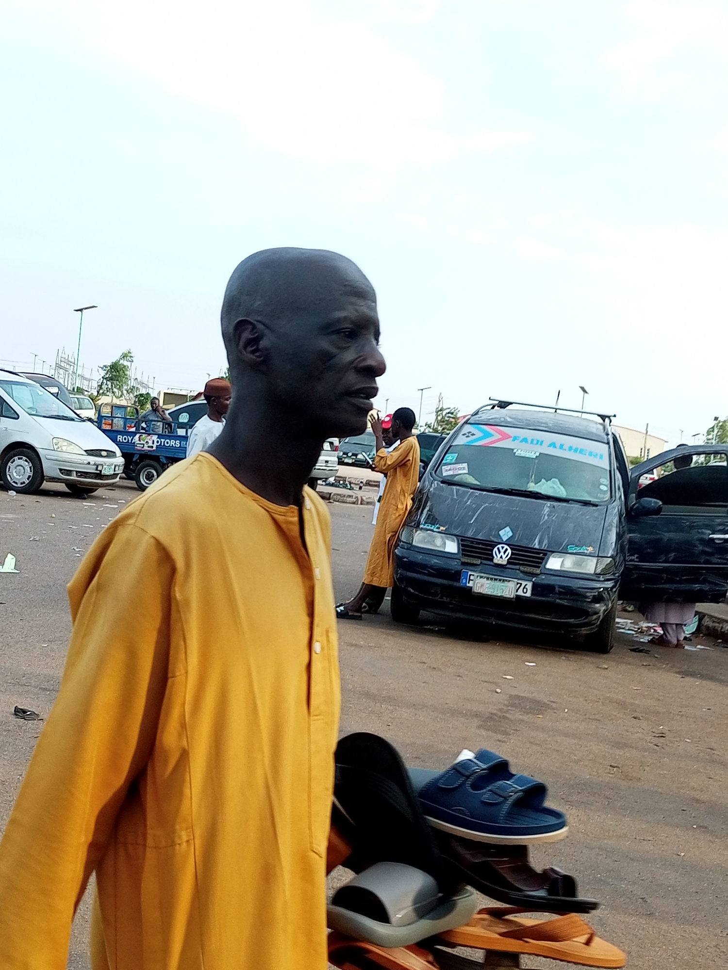 In Gombe, Nigeria, a man with a shaved head and dark skin is walking away from the camera, wearing a bright yellow garment. He is in a parking lot filled with cars and other people. The man in the foreground is the primary subject. In the background, a black minivan with a sticker that reads "FADI ALHERI" is parked with its doors open. Another man in a yellow outfit is standing near the minivan. A small blue truck with "ROYAL MOTORS" written on it is also visible, along with a silver car. The ground is paved asphalt. The sky is overcast.