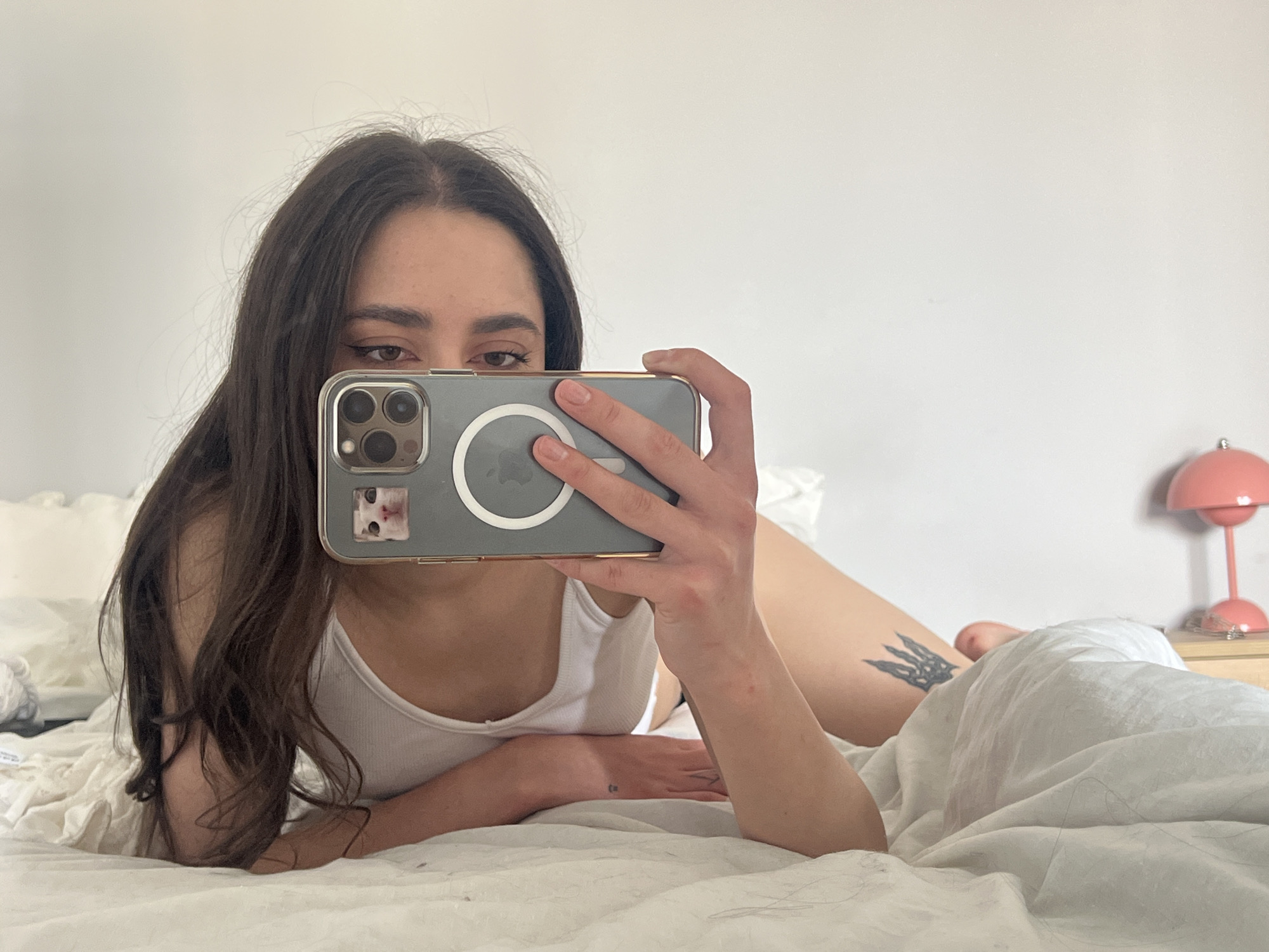 A woman is lying in bed taking a selfie. She is wearing a white tank top. Part of her face is obscured by the phone she's using. The phone is gray and has a clear case. She has brown hair and is wearing eyeliner. There is a tattoo on her leg, and another small tattoo on her hand. The bed has white sheets. A pink lamp sits on a bedside table. There is no clear indication that this image was taken in Ternopil, Ukraine.