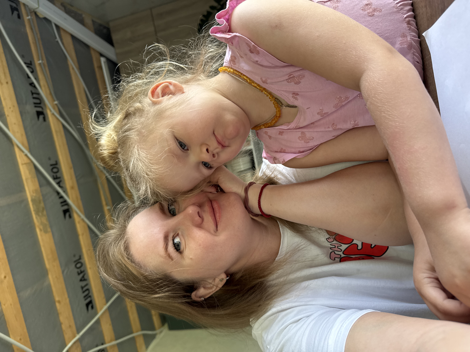 A woman and a young child are posing for a selfie. The woman has fair skin, blonde hair, and blue eyes, and is wearing a white t-shirt. The child, with blonde hair, blue eyes, and a pink dress, is nestled against her, close to the woman's face. The background shows a wall with visible insulation materials marked "JUTAFOL" attached to wood slats. The scene likely takes place indoors in a location near Ферма, Russia.