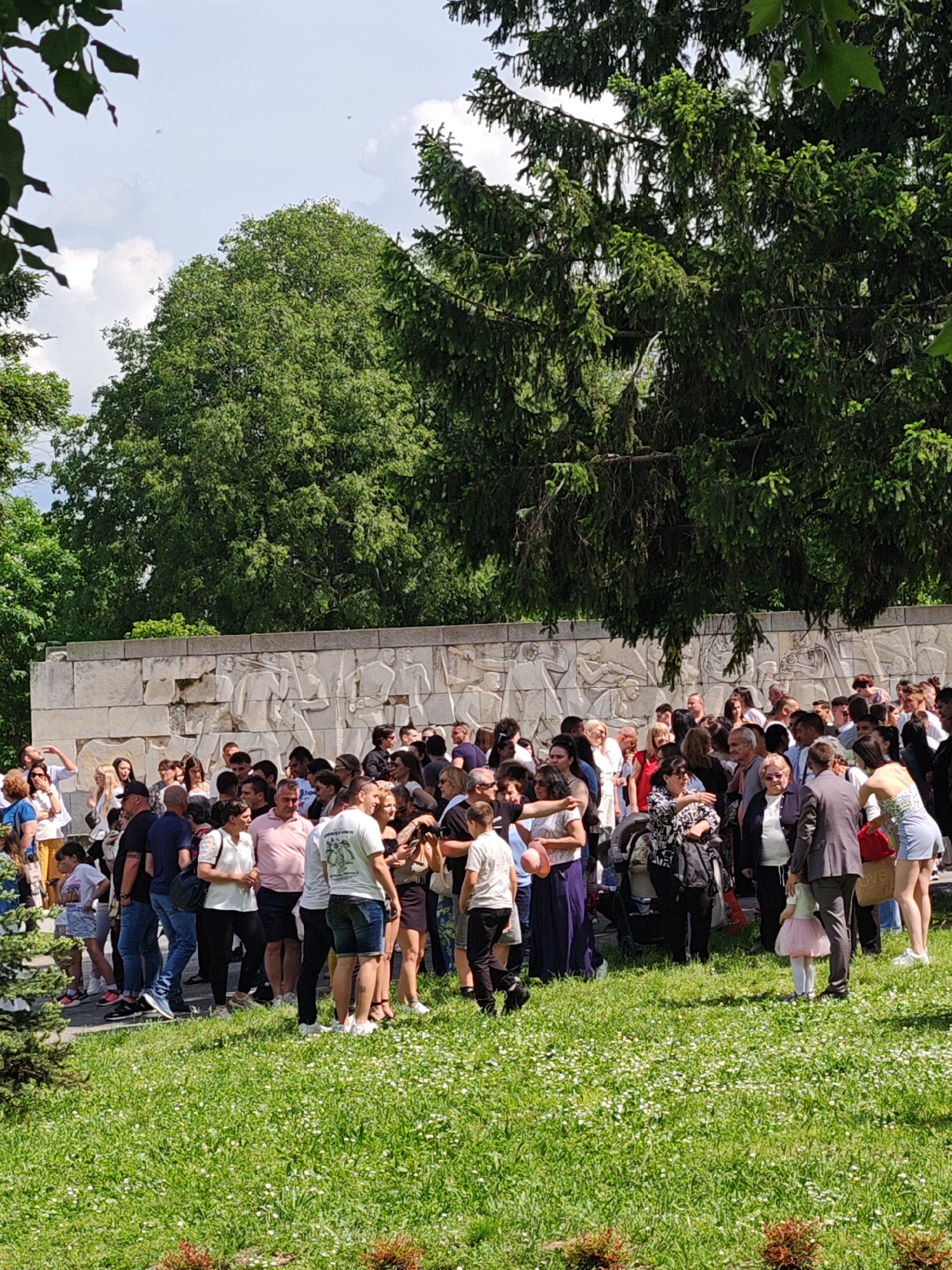 A large group of people stands on a grassy area with white flowers in front of a long, stone memorial wall depicting figures in relief. The location is in the city of Shumen, Bulgaria. The crowd is diverse in age and attire, with some individuals in casual clothing like shorts and t-shirts, while others are more formally dressed. Some people are interacting in small groups, talking and hugging, while others appear to be observing something. A few children are present. The background consists of trees and a partially cloudy sky.