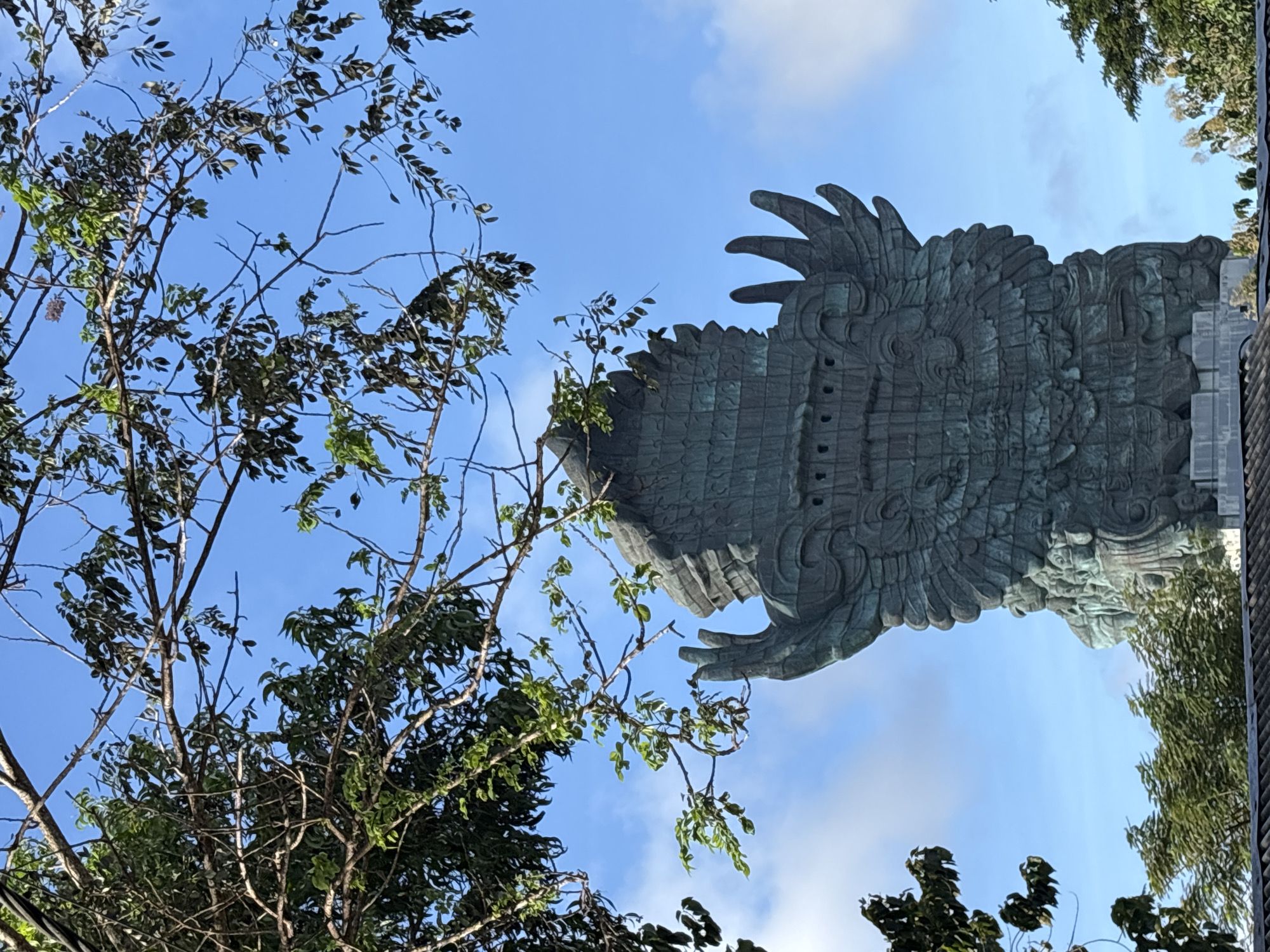 A low-angle shot shows part of the Garuda Wisnu Kencana statue in Kuta Selatan, Indonesia. The statue appears in the upper right of the image, extending down the right side. The statue, appearing in a teal color, is highly detailed and shows the large, feathery wings of a mythical bird. A tree with sparse foliage frames the statue on the left side of the image. In the space between the statue and the tree, a bright blue sky is visible. Other trees are also at the bottom.