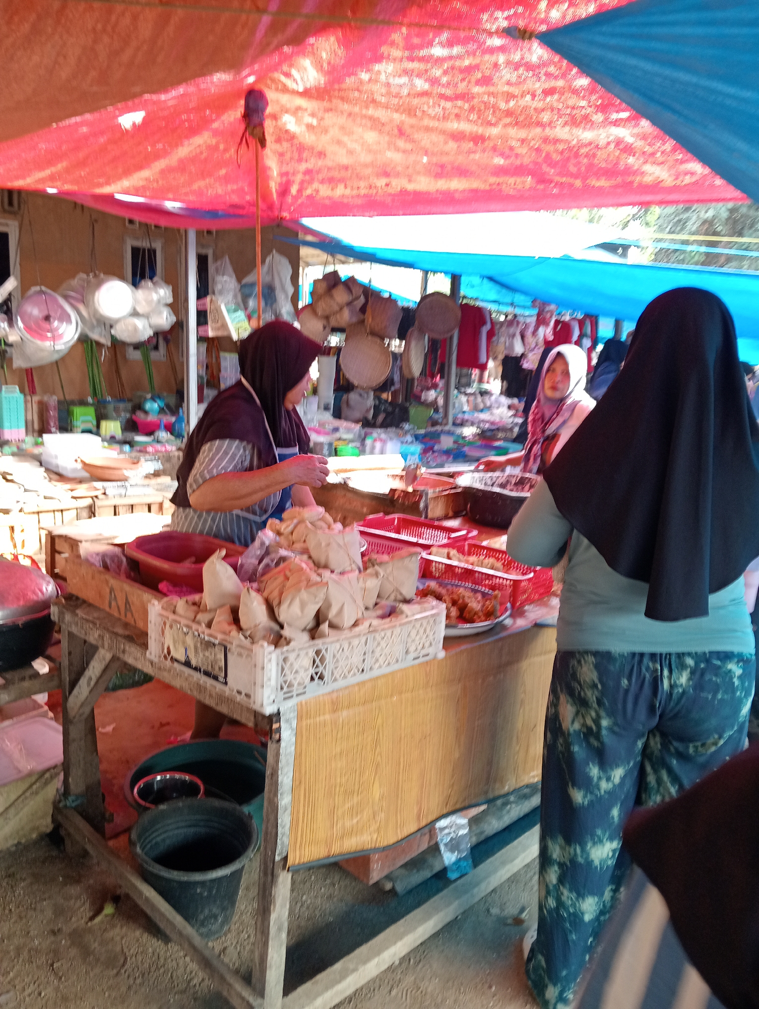 An outdoor market scene in Gunung Sugih, Indonesia, under a red and blue tarp shelter. Three individuals are visible. In the foreground, a person wearing a black hijab and tie-dye patterned pants faces away from the camera, looking towards a market stall. To their left, a vendor, wearing a brown hijab and a striped apron over a gray and white striped shirt, is engaged in an activity at the stall, possibly serving food. In the background, another person wearing a light pink hijab and a patterned scarf is partially visible, also near a market stall. The stall displays various items, including what appears to be food wrapped in brown paper in white plastic crates, and several red plastic baskets containing food items. To the left of the stall, household items such as pots and pans are displayed. The ground appears to be dirt or gravel. Various buckets are situated at the base of the wooden market stall.