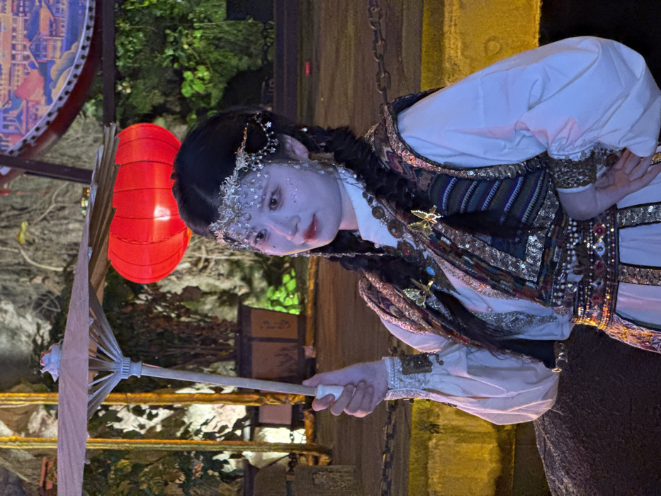 A woman in an elaborate costume stands under a red lantern. Her attire includes a white collared shirt, a patterned vest, and ornate jewelry adorning her head and wrists. She holds a white parasol. Her face is painted with shimmering accents. The scene is dimly lit, with foliage and building structures visible in the background.
