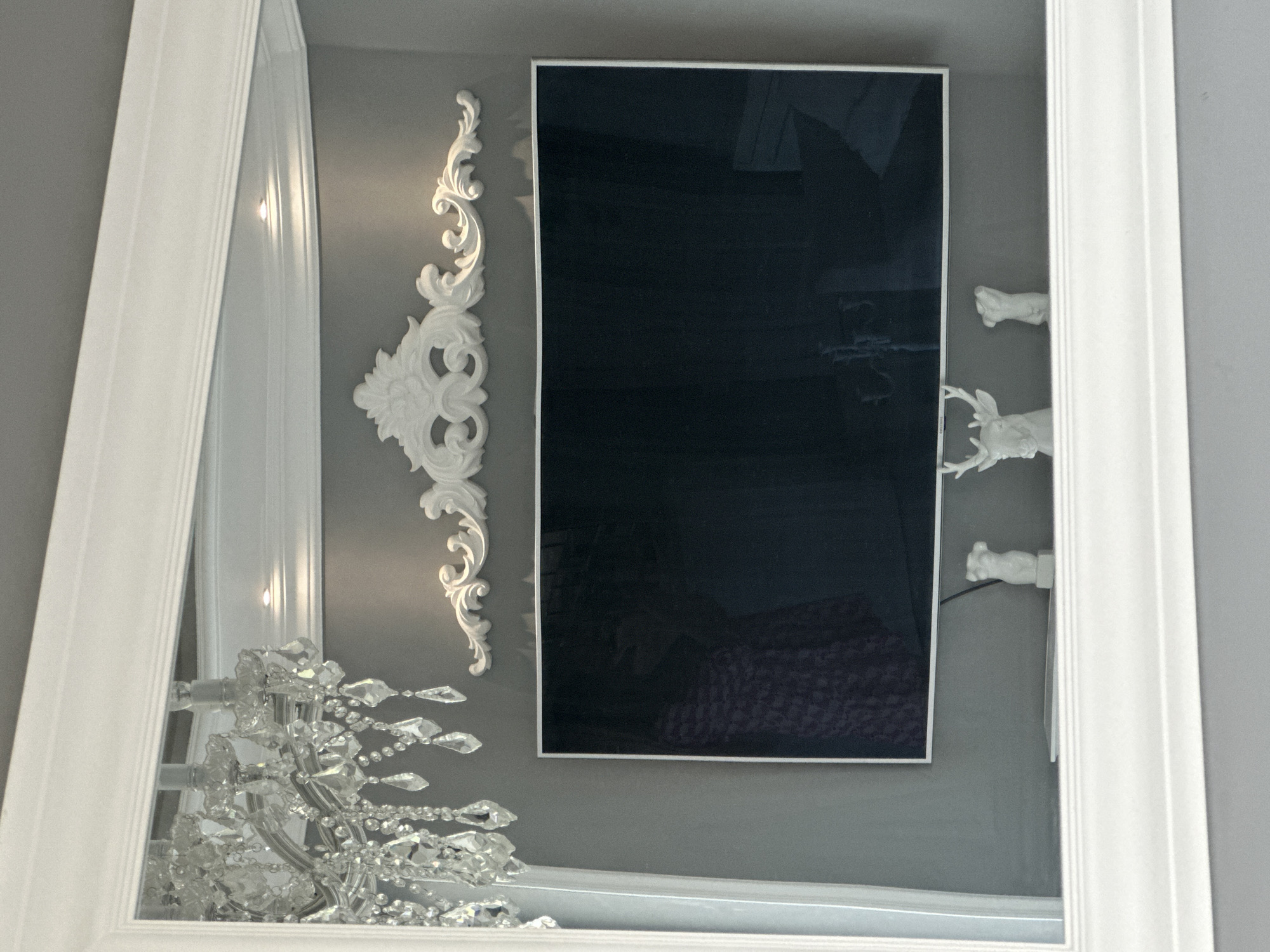 The image displays a detailed reflection within a large, white, ornate mirror of a section of an interior room. The reflected scene features a smooth, medium-grey wall serving as the backdrop. A white crown molding or cornice runs along the top of the wall, with multiple small, bright recessed spotlights embedded within it, casting light downwards.
Centered on the grey wall, above a television, is a large, white, three-dimensional decorative wall ornament. This piece exhibits classical baroque or rococo-style scrolling patterns, culminating in a prominent central floral or foliate motif. Below this ornament, a large flat-screen television with a slim white bezel is mounted. The television screen is dark and highly reflective. Visible reflections in the screen include a person wearing a garment with a dark purple or maroon pattern on the left side, and faint outlines of what appear to be furniture or another room on the right side.
Beneath the television, on a partially visible surface, three white decorative figurines are arranged. From left to right, these include a small, abstract statue resembling a classical human torso, followed by a larger sculpture of a deer head with prominent antlers, and then another small, abstract human torso statue. A flat, white electronic device, possibly a tablet or media player, rests on the surface between the leftmost torso statue and the deer head. A black cable is visible behind the deer head, likely connected to the television or the electronic device.
In the upper left portion of the mirror's reflection, a multi-tiered crystal chandelier is partially visible. The chandelier is adorned with numerous clear crystal drops and facets. The light sources within the chandelier appear to be unlit. The prominent white, decorative frame of the mirror itself occupies the foreground, framing the entire reflected scene.