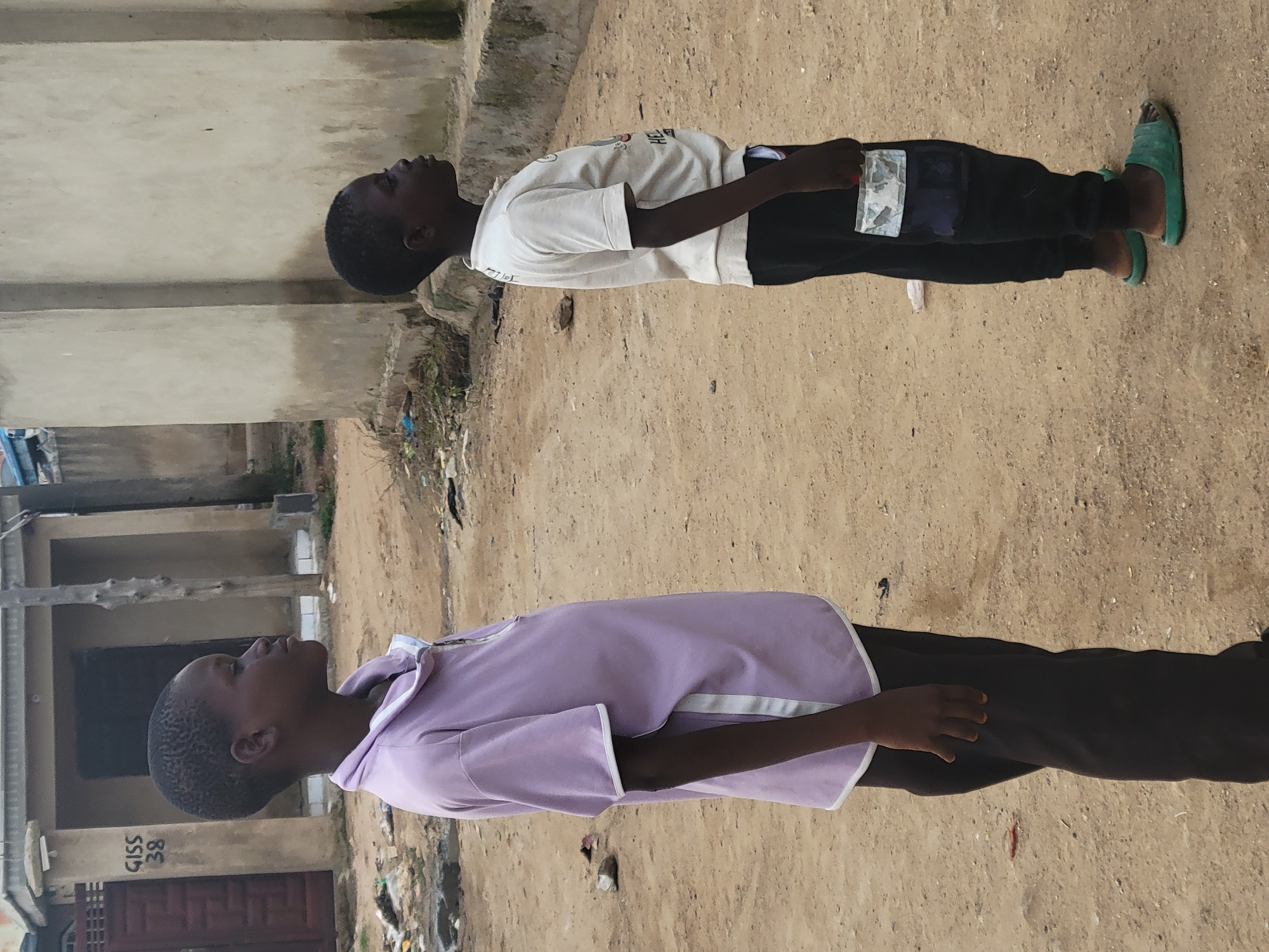 In Madalla, Nigeria, two young boys stand in profile facing right on a dusty, unpaved surface. The boy on the left, wearing a light purple zipped polo shirt and dark pants, has short hair. The boy on the right is in a white t-shirt with a graphic and black pants, also with short hair. His right hand is in his pocket, holding what appears to be paper currency, possibly Nigerian Naira. Both boys are wearing open-toed sandals. The background shows a building with a textured concrete wall and a doorway. The ground is uneven and scattered with debris.