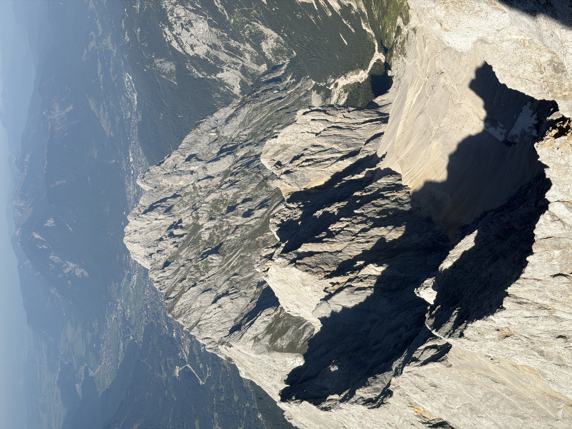This aerial photograph, taken from a high vantage point in the Bavarian Alps near Garmisch-Partenkirchen, Germany, offers a panoramic view of rugged mountainous terrain and a valley settlement. The foreground is dominated by the stark, grey, and rocky peaks of a mountain range, with sharp ridges and steep inclines casting long shadows in the bright sunlight. The texture of the rock faces is detailed, showing layers and fissures.
In the mid-ground, a prominent mountain peak rises, its ridgeline appearing serrated and imposing. Beyond this, the landscape transitions into a verdant valley. A town, likely Garmisch-Partenkirchen, is visible nestled within this valley, characterized by a dense cluster of buildings with red rooftops and scattered smaller structures. Roads and paths snake through the urban area and into the surrounding green fields and forests.
Further into the background, successive layers of mountains recede, their forms softened by atmospheric haze, creating a sense of depth and vastness. The sky is a clear, pale blue, indicative of fair weather. The image captures a dramatic juxtaposition of the raw, untamed nature of the mountains with the organized, human-made environment of the town below. There are no people or discernible actions visible in the frame.