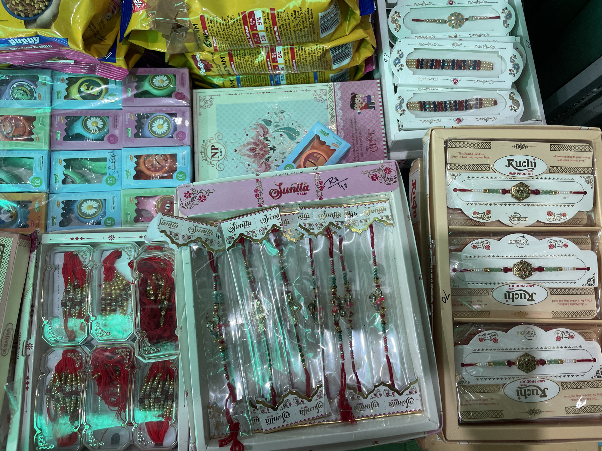 A retail store in Bhagsu, India, displays various gift items and packaged goods on shelves. The foreground shows several boxes of rakhis, a traditional Hindu bracelet. To the left, a beige box contains individually packaged rakhis with red strings and decorative beads. In the center, a white box is filled with multiple rows of rakhis, each enclosed in clear plastic packaging. These rakhis also feature red strings and a variety of colorful beads and embellishments. To the right, smaller, brightly colored boxes are stacked, some with images of cartoon characters and flowers. A large, yellow bag of food product with nutritional information printed on it is visible on the far right. The overall lighting suggests an indoor environment, possibly a market or shop.