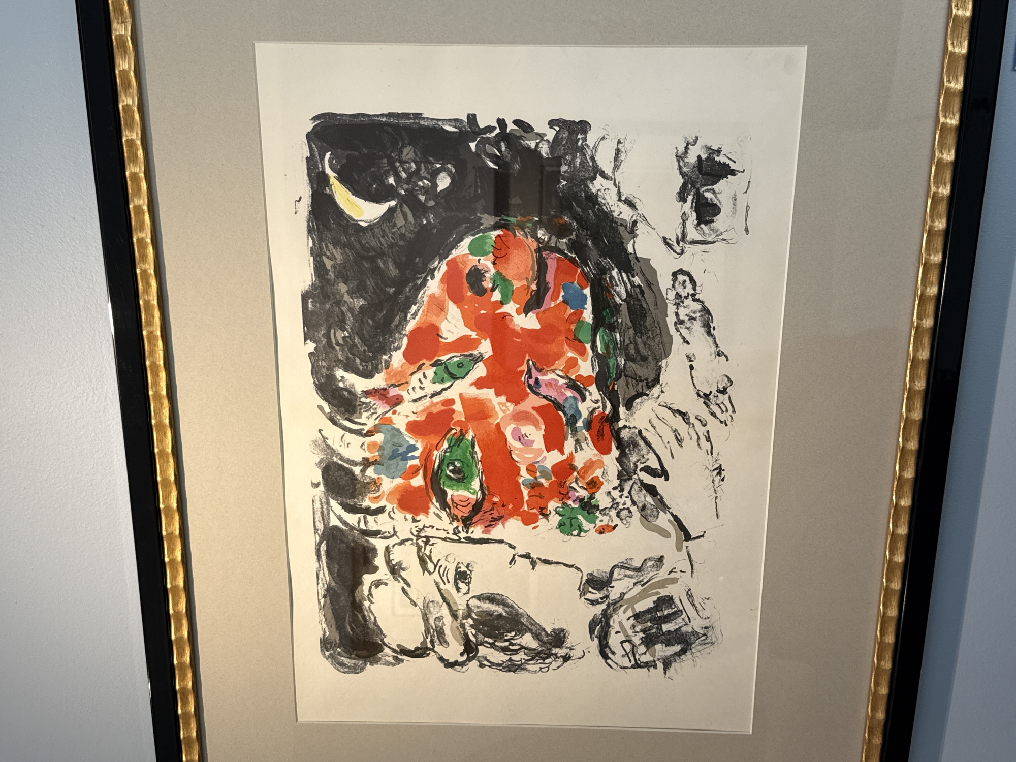This is a photograph of a framed artwork, possibly a lithograph or print, hanging on a wall. The artwork itself is a colorful and abstract depiction of a face or faces, rendered in black, white, red, green, blue, and yellow. The style is reminiscent of Surrealism, with distorted features and overlapping elements. The prominent element is a large, orange-red form with striking green eyes. Black ink lines define facial contours and abstract shapes. To the left of the central image, there are suggestions of profile faces and fragmented forms in black and white. To the upper right, a yellow crescent moon is visible against a dark, abstract background. The artwork is matted with a pale beige mat and enclosed in a black frame with a gold, textured inlay along the inner edge. The wall behind the artwork is a light grey, and the wall below the frame is painted a light blue. The photograph is taken from a slight angle, looking up at the artwork. The location is not specified in the image or provided context.