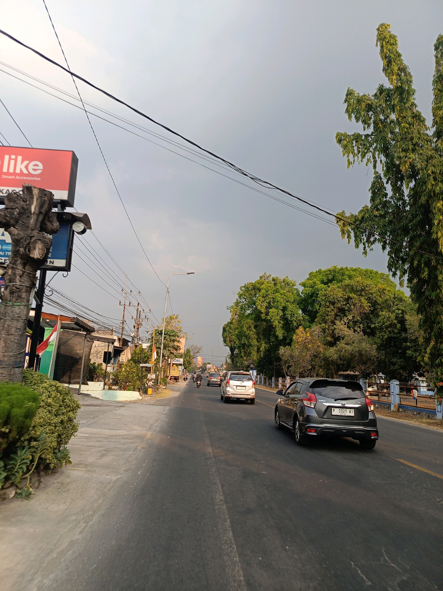 The image depicts a street scene in Kincang Wetan, Indonesia, under a gray, overcast sky. The perspective is from a vehicle traveling on the right side of the road. On the left side of the road, a red billboard with white text reads "like" and "Smart Accessories" is visible, mounted on a weathered wooden pole that appears to be a tree trunk. Further down the left side, there are shops and vegetation, including a green and white awning.
The road itself is asphalt and has lane markings. Several vehicles are present. A dark gray hatchback is in the foreground on the right lane, facing away from the camera, with its license plate reading "P 1529 WT". A silver hatchback is ahead of it in the same lane. In the distance, other vehicles, including motorcycles, are visible on the road.
On the right side of the road, tall, slender trees with dense green foliage line the street, with a blue fence visible behind them. The environment appears to be a moderately busy urban or suburban area with overhead power lines crisscrossing the sky. The overall lighting suggests it might be late afternoon or early evening, with the sun not directly visible due to the cloud cover.