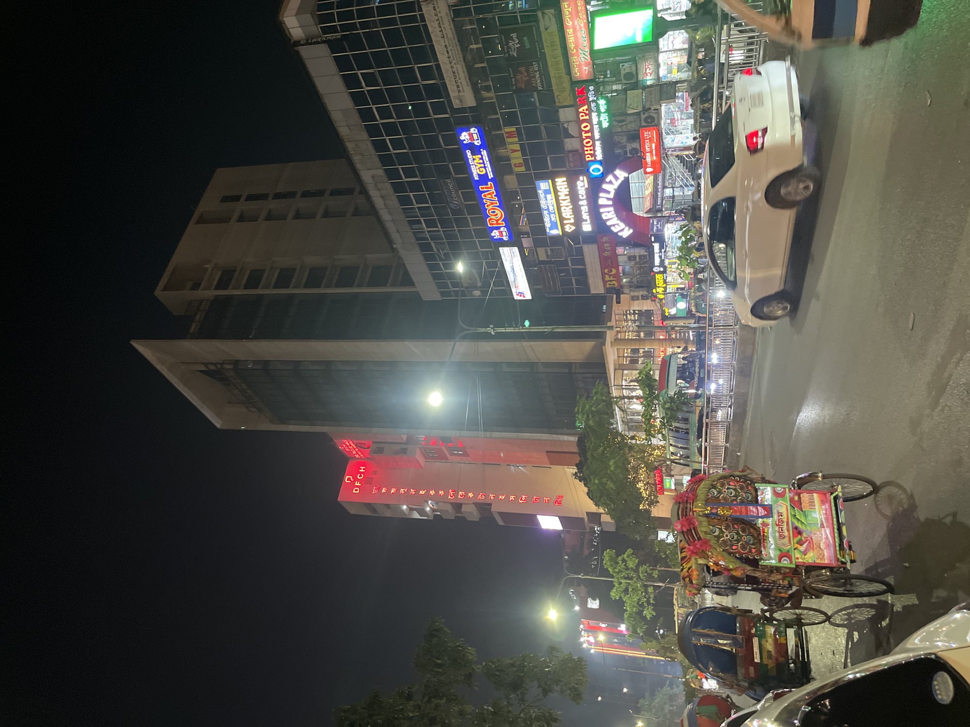 The image portrays a nighttime street scene in Dhaka, Bangladesh. Illuminated signs for "Royal Gym," "Photo Park," and "Keari Plaza" are visible on buildings, indicating a commercial area. A white car is parked on the side of the road. A vividly decorated rickshaw is prominent in the foreground, suggesting local transportation. Other vehicles and pedestrians are present on the street, contributing to the dynamic urban environment. Tall buildings line the street, interspersed with trees. The overall atmosphere is vibrant, typical of a bustling city at night.