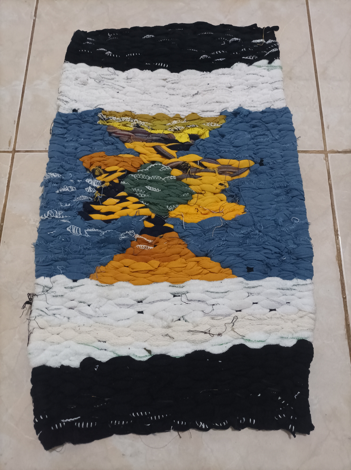 The image displays a rectangular woven rug, likely a doormat or small accent rug, lying flat on a tiled floor. The rug is vertically oriented and features a design that appears to represent a landscape. The top and bottom sections of the rug are solid black with horizontal white stitching. Above the black section, there is a wide band of white, followed by a section of blue fabric representing water or sky. The central portion of the rug depicts a tree or mountain-like form with yellow, orange, and green fabric elements, suggesting foliage or rocky terrain. The rug appears to be handmade, with visible textured fabric and loose threads at the edges. The floor is composed of square, beige tiles with grout lines. The lighting suggests an indoor environment, possibly a home. There is no indication of the city of Nganjuk, Indonesia, or any specific urban context within the image itself.