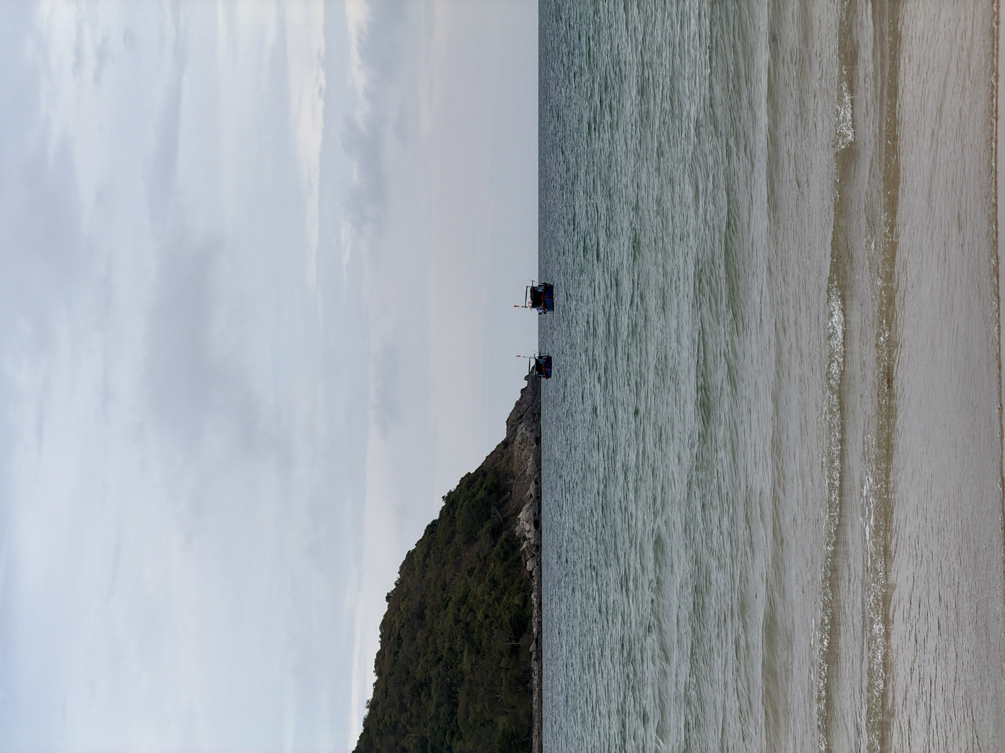 The image shows a beach in Cam Lam, Vietnam. The scene is split vertically, with the sky and land on the left and the sea on the right. The sky is overcast. On the land, there is a mountain with dense green trees. Two fishing boats are visible on the sea. The sea is calm, with gentle waves reaching the shore. The beach is sandy and appears to be wet.