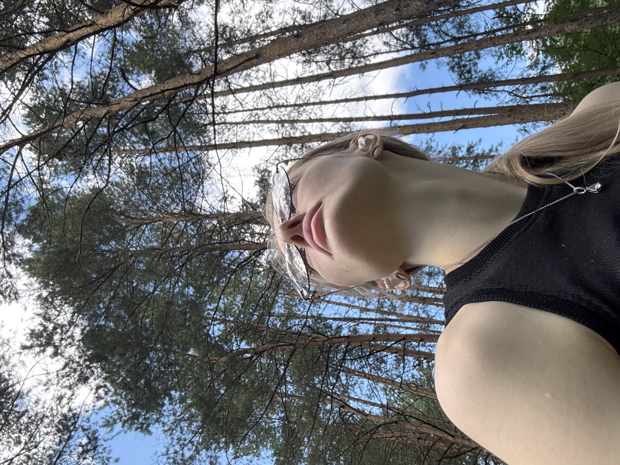 The image shows a low-angle shot of a young woman wearing glasses and a black top, looking up towards the sky. She has blonde hair and is wearing earrings and a necklace. The background is a forest with tall pine trees and a blue sky with some clouds visible. The photo appears to be taken outdoors during the daytime. The location is not specified in the image.