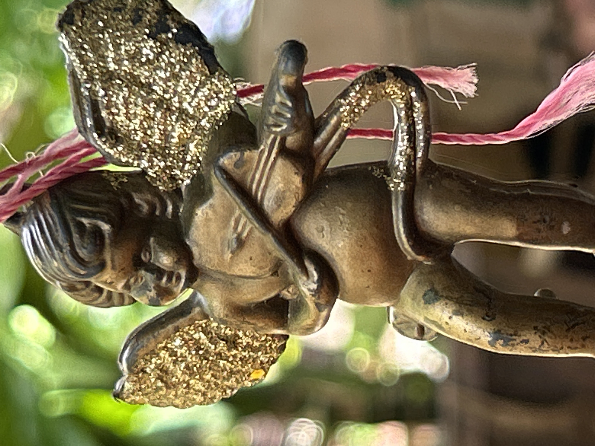 An antique, bronze-colored cherub figurine, adorned with gold glitter on its wings and tail, is depicted holding a miniature violin. The cherub is suspended by a pink braided string. The background consists of out-of-focus green foliage, suggesting an outdoor setting. The figurine appears to be a decorative item, possibly a garden ornament or a charm. The image does not provide specific information about its location in Santa Ana, Venezuela.