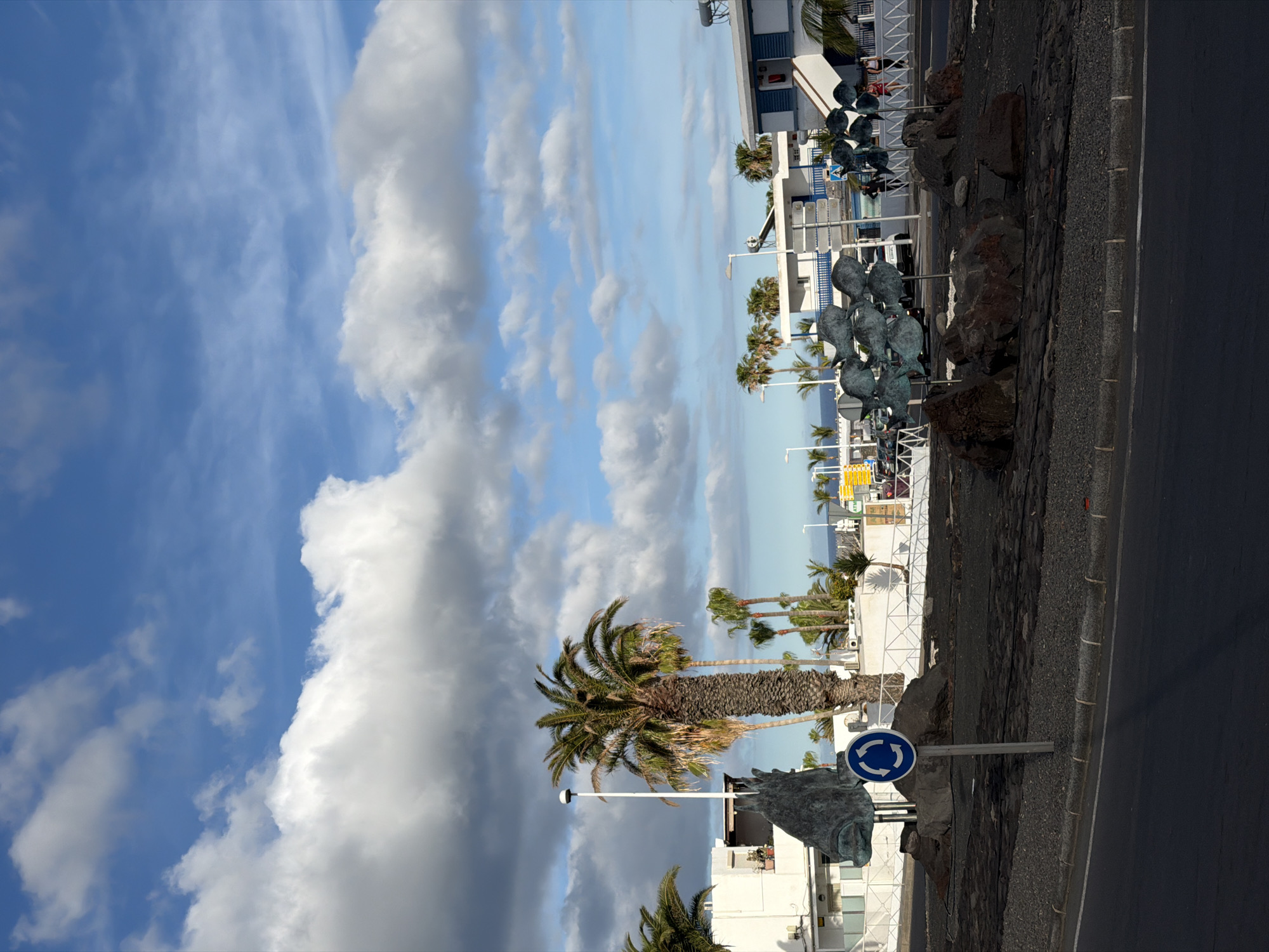 The image captures a street scene in Tías, Spain. The sky is filled with scattered white clouds against a blue backdrop. Palm trees line the street, and a sculpture of a horse head is visible, adjacent to a roundabout sign. Statues resembling fish are placed along the roadside. Buildings are present in the background, and the photograph is taken at ground level.