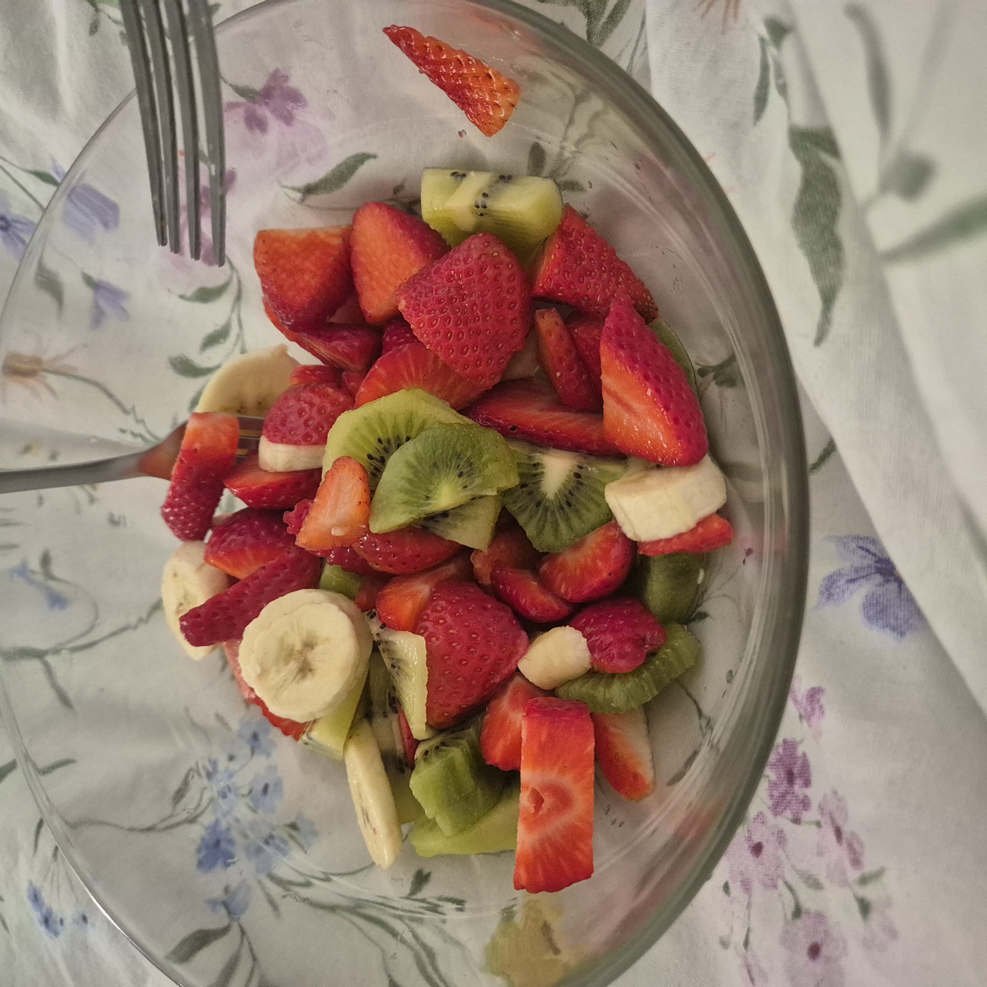 A top-down shot reveals a clear glass bowl filled with a colorful fruit salad, composed of sliced strawberries, kiwi, and bananas. A fork pierces the salad. The bowl sits on a floral-patterned fabric. The location is unknown.