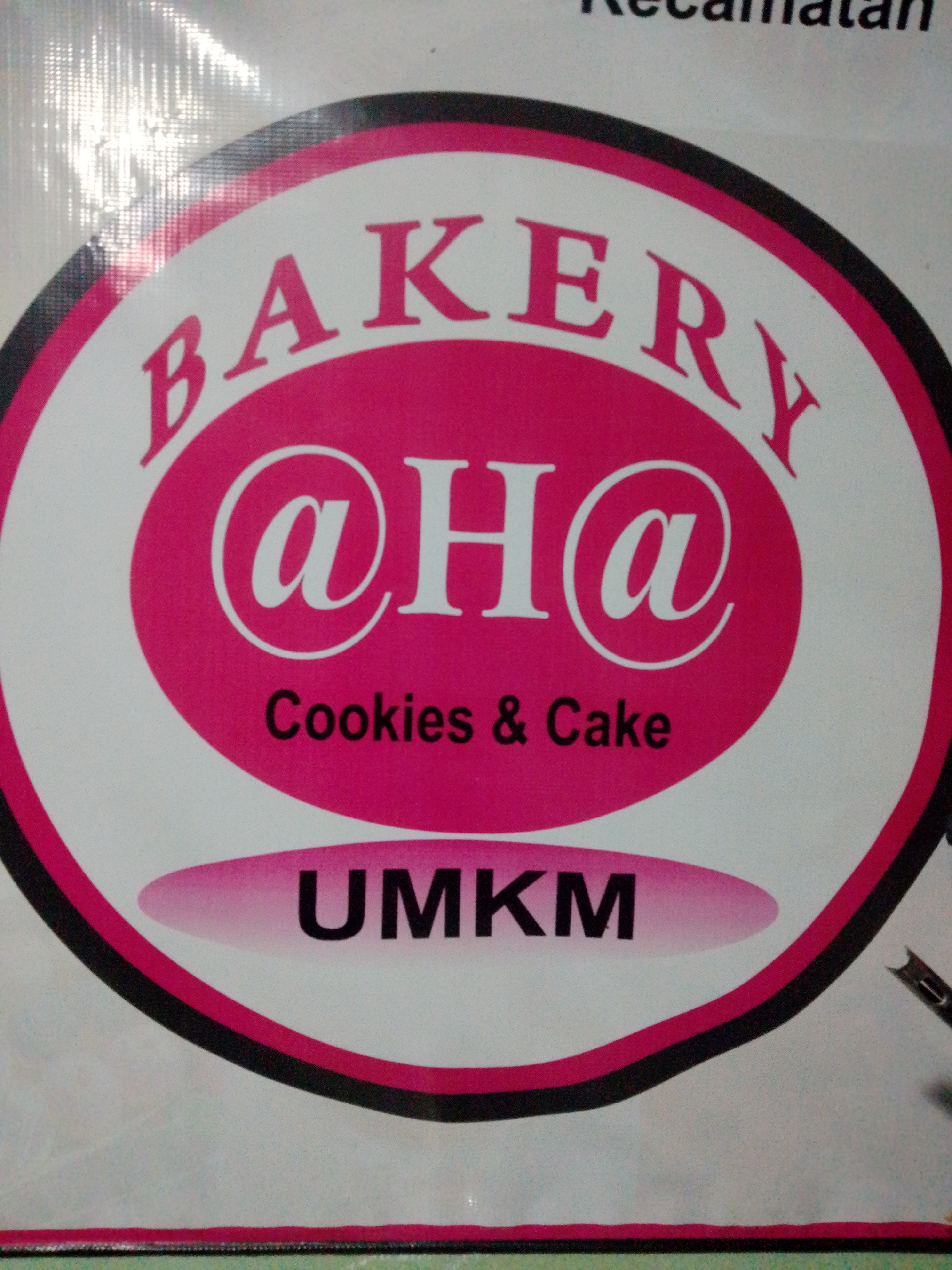 A close-up of a sign for "BAKERY @ H@ Cookies & Cake UMKM" is shown. The sign is oval-shaped with a hot pink border and a black inner border. Inside the pink border, the text "BAKERY" is written in large, white, stylized letters that curve along the top. In the center, "@ H@" is written in white, with the "@" symbols in a circular font. Below this, "Cookies & Cake" is written in smaller, black text. At the bottom, the word "UMKM" is written in large, black, sans-serif letters. The sign appears to be made of vinyl or a similar material and is likely displayed outdoors, possibly on a shop front or a banner. The background of the image is a slightly blurred, off-white surface with a textured pattern. The lighting suggests an indoor or shaded outdoor setting. There are no people or specific environmental features visible beyond the sign itself. The text "UMKM" is an acronym for "Usaha Mikro, Kecil, dan Menengah," which translates to "Micro, Small, and Medium Enterprises" in Indonesia, indicating this bakery is a small business. This type of signage is commonly found in local businesses throughout Indonesian cities, including Bandar Lampung, to advertise products and services.