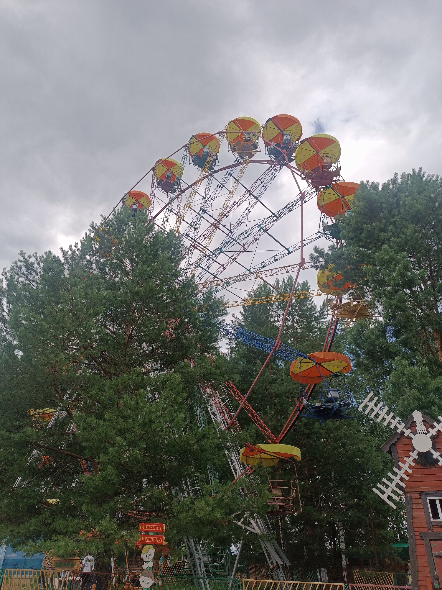An amusement park ride, possibly a Ferris wheel, is visible amidst trees under a cloudy sky. Several gondolas, colored orange and yellow, are attached to the circular structure. In the foreground, a small wooden structure resembling a windmill is present, and further left, a fence surrounds part of the park. Two signs with cartoonish faces and text, possibly in Cyrillic script, are also visible. The general atmosphere suggests a recreational area.