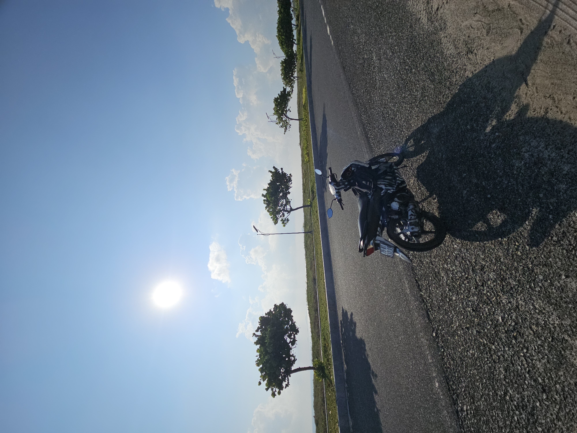 The image depicts a sunny day in Bac Cam Ranh Ward, Vietnam. A motorcycle is parked on the side of a road, casting a long shadow. Trees line the roadside, separating the road from grassy fields. The sky is mostly clear with scattered clouds. The sun is visible in the lower left corner of the frame.
