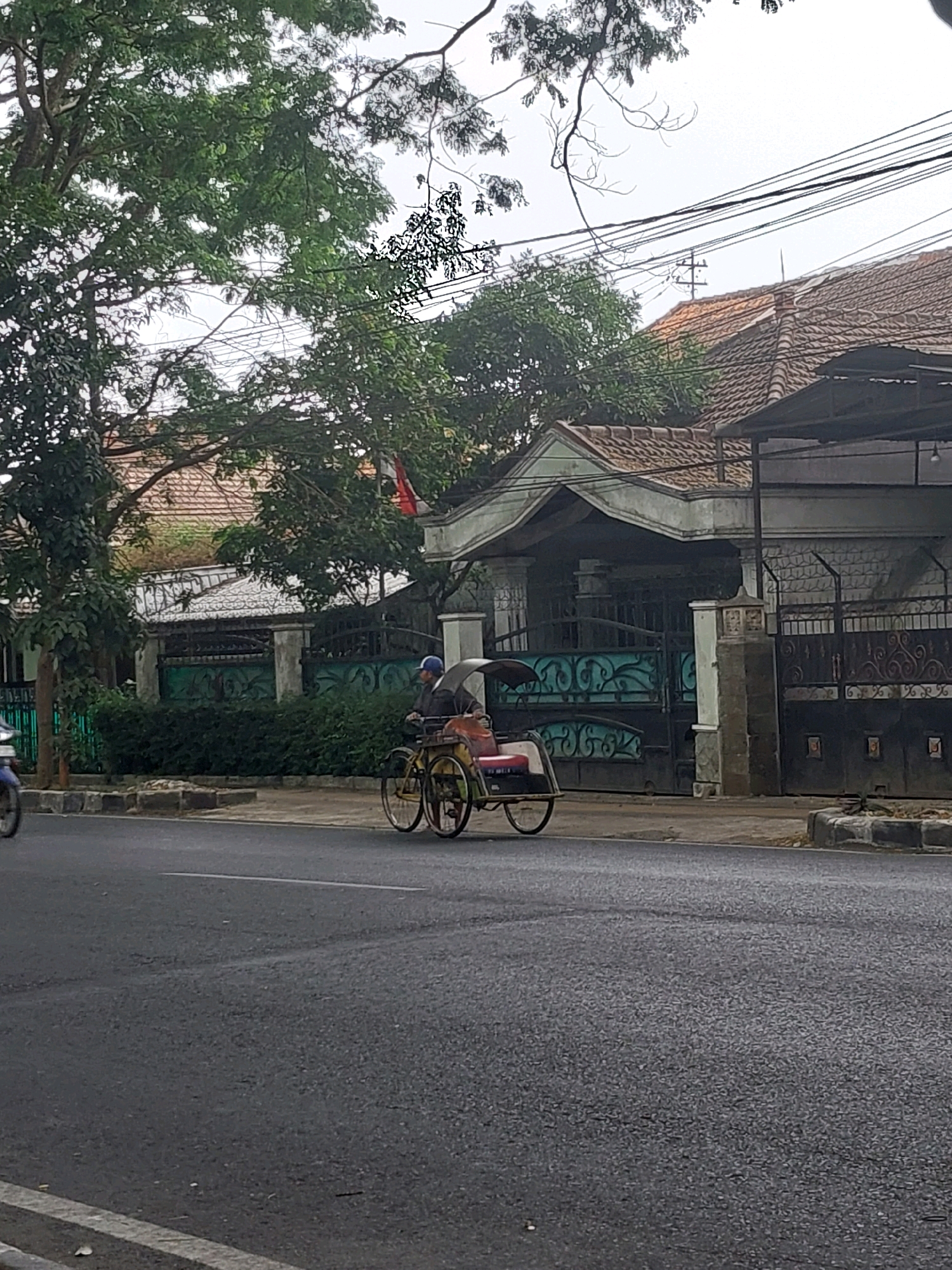 A becak driver pedals a yellow becak along a paved street in Malang, Indonesia. The driver wears a blue cap. The becak has a canopy and a red seat. Behind the becak, a residential fence is visible, with a house and trees in the background. The sky is overcast.