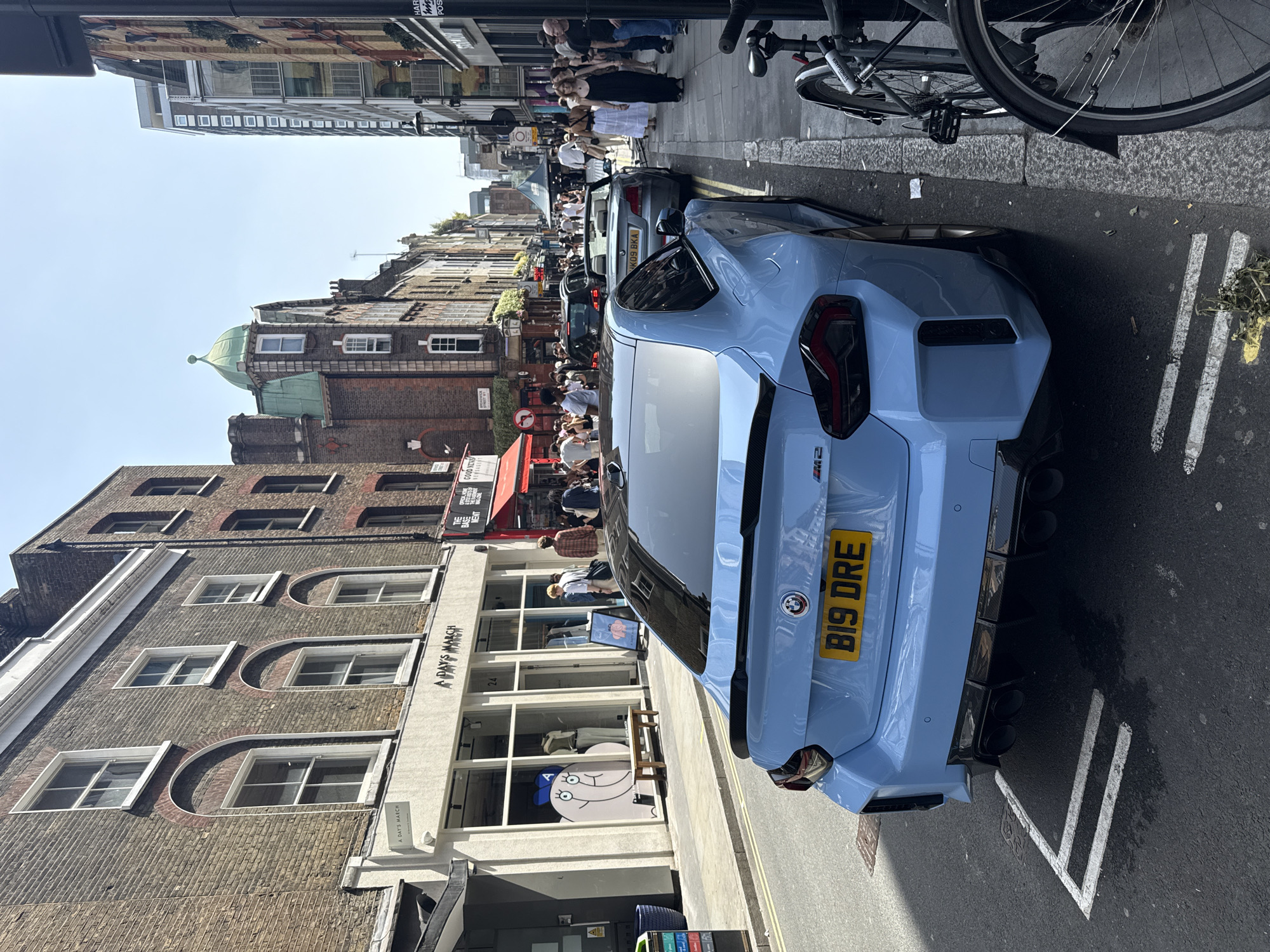 A street scene in London, United Kingdom, shows a powder blue BMW M2 parked on the side of the road. The car has the license plate "B19 DRE." To the left of the car is a building with a storefront labeled "A DAY'S MARCH." People are visible on the sidewalk near the shop. Farther down the street, a red awning is visible over another shop with the sign "GOOD NEWS." Beyond that, more people are walking on the sidewalk. In the background, there is a building with a green dome on top. The street appears busy with pedestrians and parked cars. A bicycle is parked on the sidewalk, next to the car. The sky is bright and clear, suggesting it is daytime.