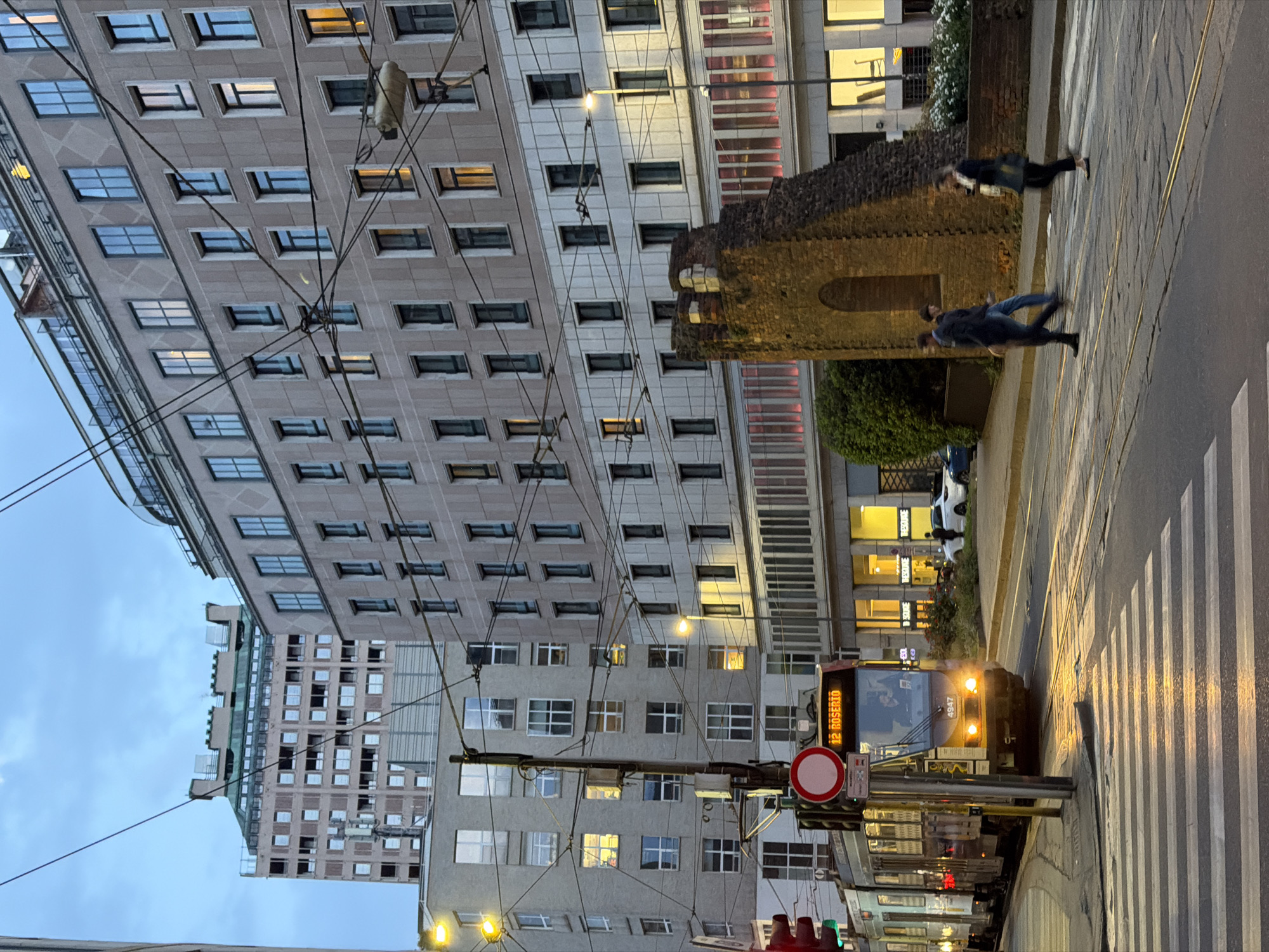 This image captures an urban street scene in Milan, Italy, at dusk or early evening. The perspective is tilted, showing tall, multi-story buildings dominating the left side of the frame, with the sky visible on the far left. Many windows in the buildings are lit, casting warm glows.
In the mid-ground, an ancient-looking stone structure with an arched opening stands, providing a historical contrast to the surrounding modern architecture. Overhead power lines crisscross the sky, indicative of tram or trolleybus infrastructure.
On the right side, a street or tram tracks are visible. A tram, bearing the number "4947" and displaying "12 ROSERIO" as its route or destination, is approaching or stopped, with its headlights brightly illuminated. Reflections of the tram's lights and surrounding illumination are visible on the wet-looking street surface. A prominent zebra crossing is marked on the pavement.
Several pedestrians are visible on the street or sidewalk near the tram, some appearing blurred due to motion. Street furniture includes a pole with a circular red and white prohibitory sign (likely "no entry" or similar) and traffic lights, with both red and green signals faintly visible. The overall atmosphere suggests a typical busy evening in a European city, characterized by the interplay of natural twilight and artificial lighting.