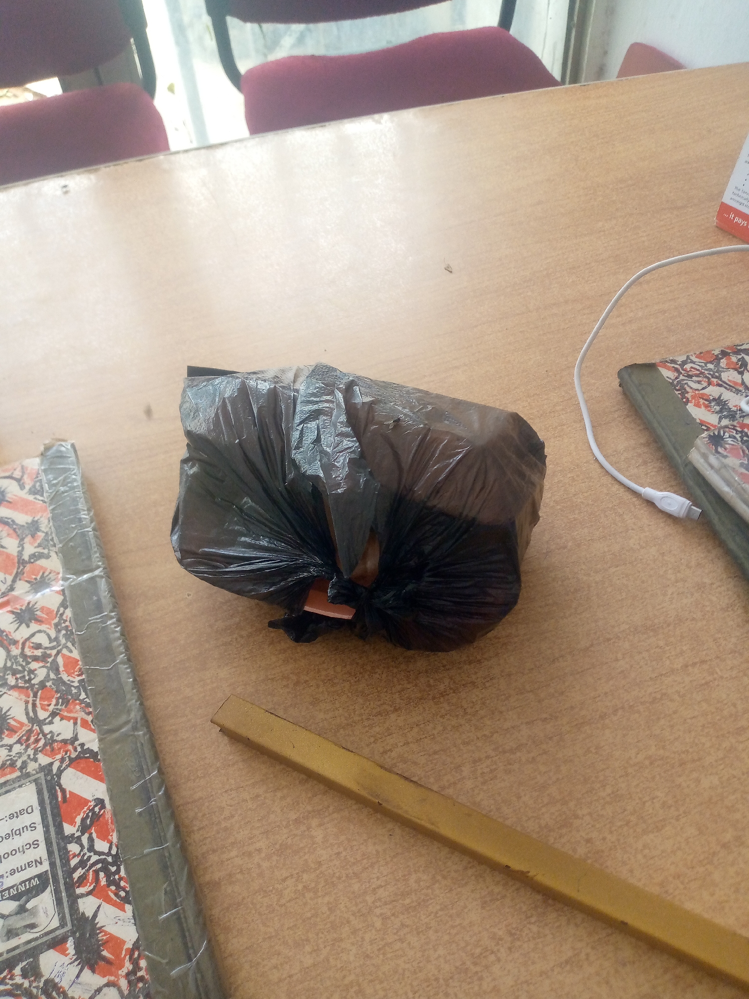 The image depicts a close-up, overhead view of a wooden desk in Jalingo, Nigeria. On the desk, a black plastic bag is centrally placed, appearing to contain some items. To the left of the bag is a notebook with a distinctive orange and black patterned cover, wrapped in clear plastic. Further to the right, another notebook, also with a patterned cover, is visible, connected to a white USB cable. A golden ruler-like object lies in the foreground, running horizontally across the lower portion of the image. The background shows out-of-focus red chairs, suggesting an indoor setting such as a classroom or office.