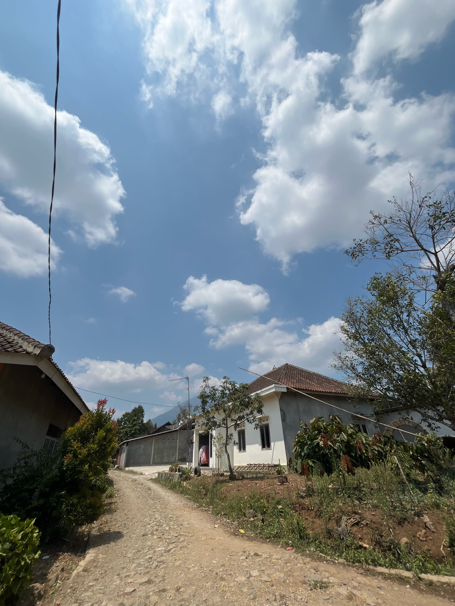 A dirt road, lined with vegetation and houses, winds through the scene. The sky above is a bright blue, dotted with fluffy white clouds. The houses appear to be simple, residential structures with tiled roofs. In the foreground, a power line hangs across the sky. The overall impression is one of a rural or semi-rural setting in the village of Margodadi, Indonesia, on a clear day. There are no people visible in the image.