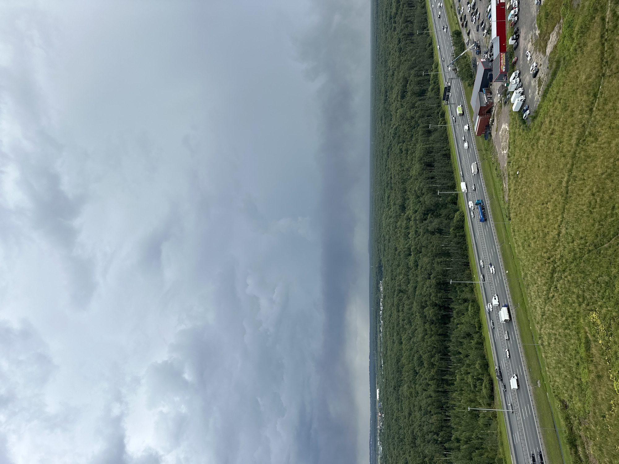 The aerial photograph shows a highway running through a dense forest and adjacent to a grassy field. Several vehicles, including cars, trucks, and a blue tanker truck, are visible on the highway. On the right side of the highway, there is a parking lot with numerous cars, and several buildings with red roofs. There are also some lamp posts along the highway and a line of trees bordering the forest. The sky is overcast with dark clouds, suggesting a stormy or rainy day. The scene appears to be in a rural or semi-rural area, given the extensive forestation. Some text is visible on the red-roofed buildings, possibly indicating businesses. The photograph is taken from a high angle, providing a broad view of the landscape and the highway activity.