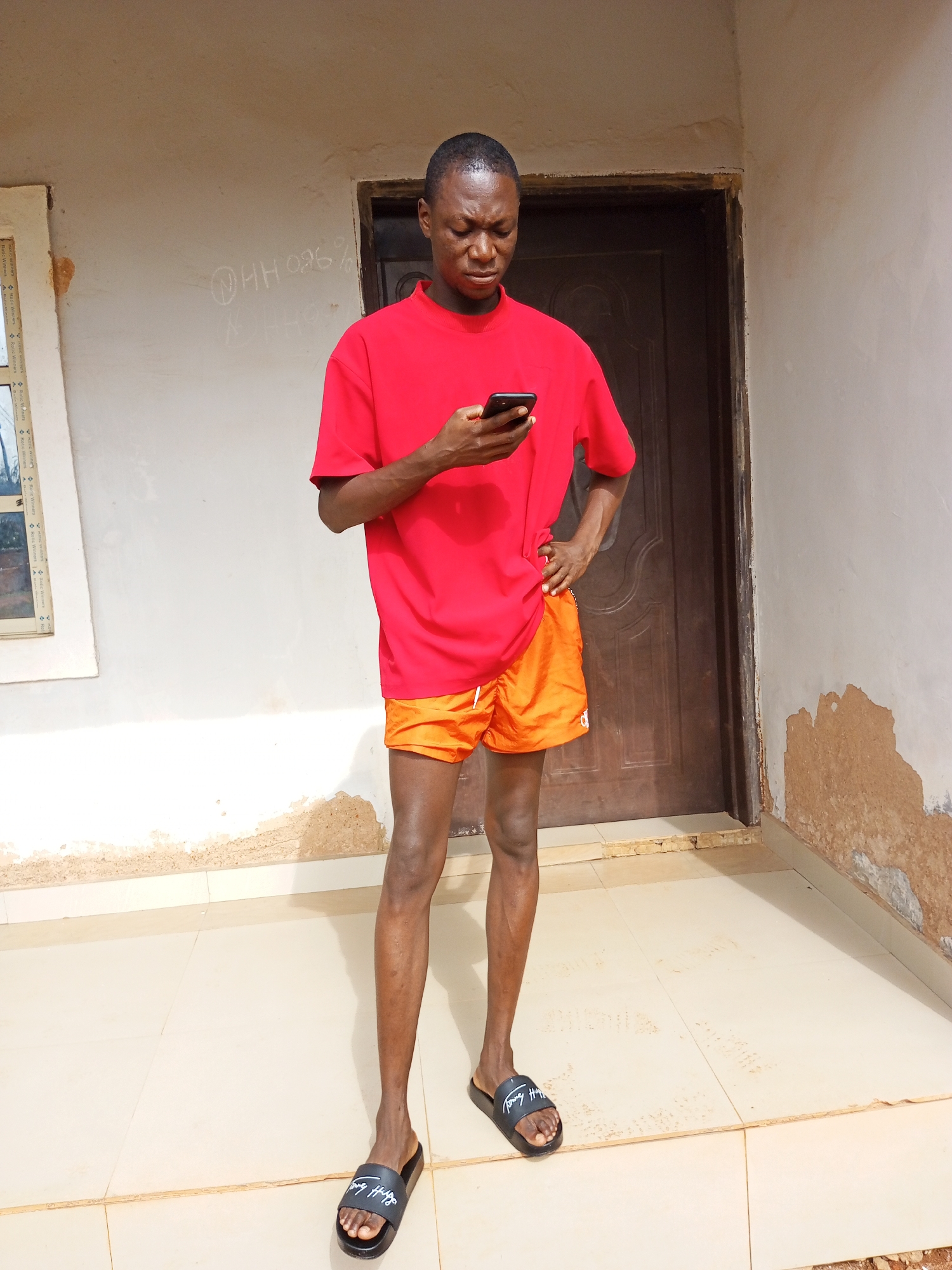 The image depicts a young man standing on a tiled porch in front of a building in Bukuru, Nigeria. The man is holding a black smartphone in his hands and looking down at it. He is wearing a short-sleeved red t-shirt and bright orange shorts. He is also wearing black slide sandals with white lettering. The man's left hand is on his hip.
The building behind him has a brown door in the center. To the left of the door is a window with white frames. The exterior of the building is painted a light color, but some of the paint is peeling off at the bottom, revealing a darker layer beneath. There is some writing on the wall to the left of the door. The porch is tiled with light-colored tiles.