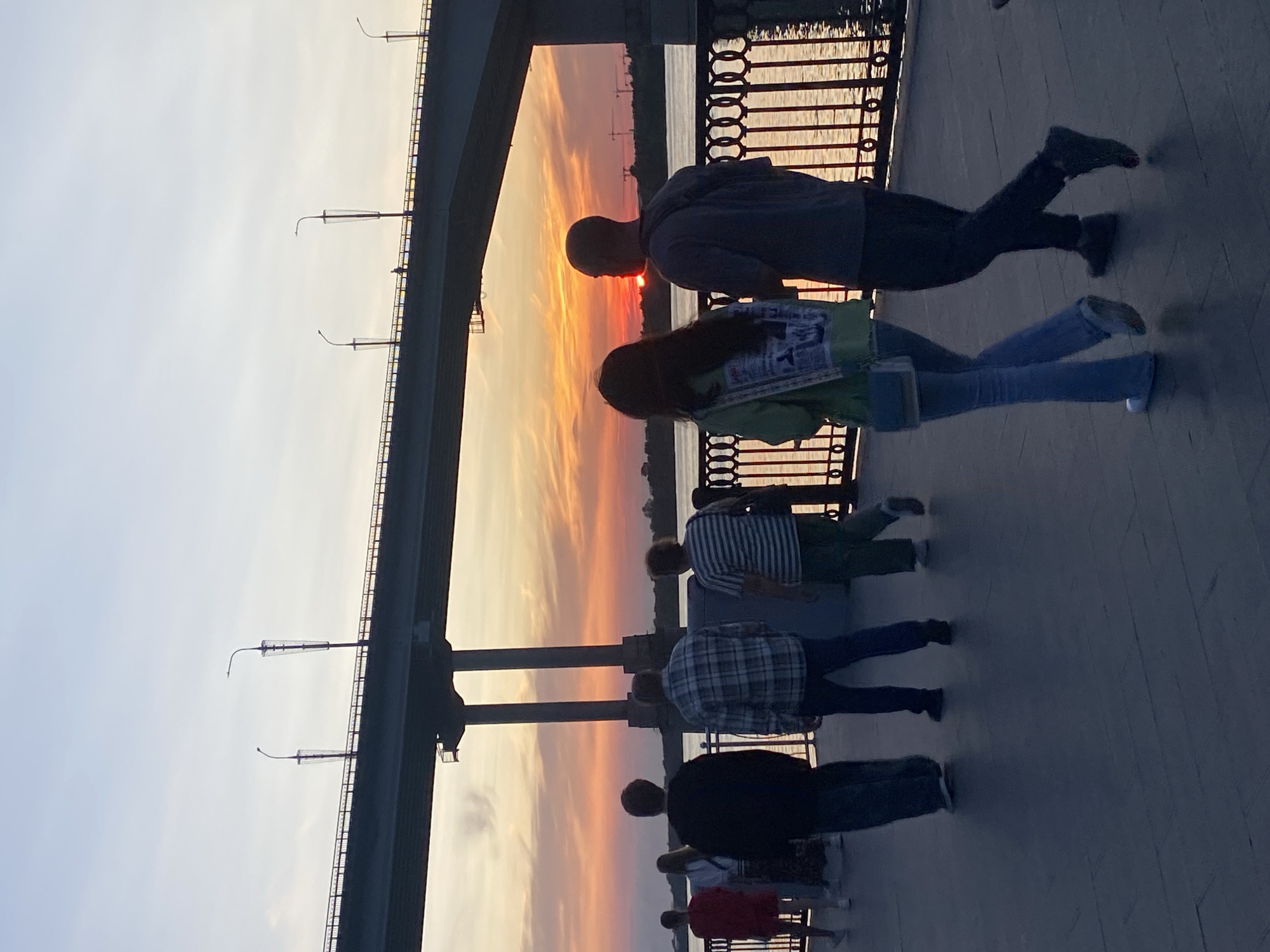 The image depicts a sunset scene over the Kama River in Perm, Russia, viewed from a promenade beneath a large bridge. Silhouetted against the vibrant orange and pink hues of the sky, several individuals are walking along the promenade. A couple is prominently featured in the foreground, facing away from the camera and towards the river. The man has a dark bag slung across his chest, and the woman wears a green jacket and light blue jeans. Further down the promenade, other figures can be seen, including a man in a plaid shirt and jeans, and a woman in a striped shirt and green pants. The promenade is paved, and a decorative metal railing separates it from the river. The bridge, a significant urban structure, spans the river in the background, with lampposts visible along its edge. The overall atmosphere is tranquil, capturing a moment of evening leisure in the city.